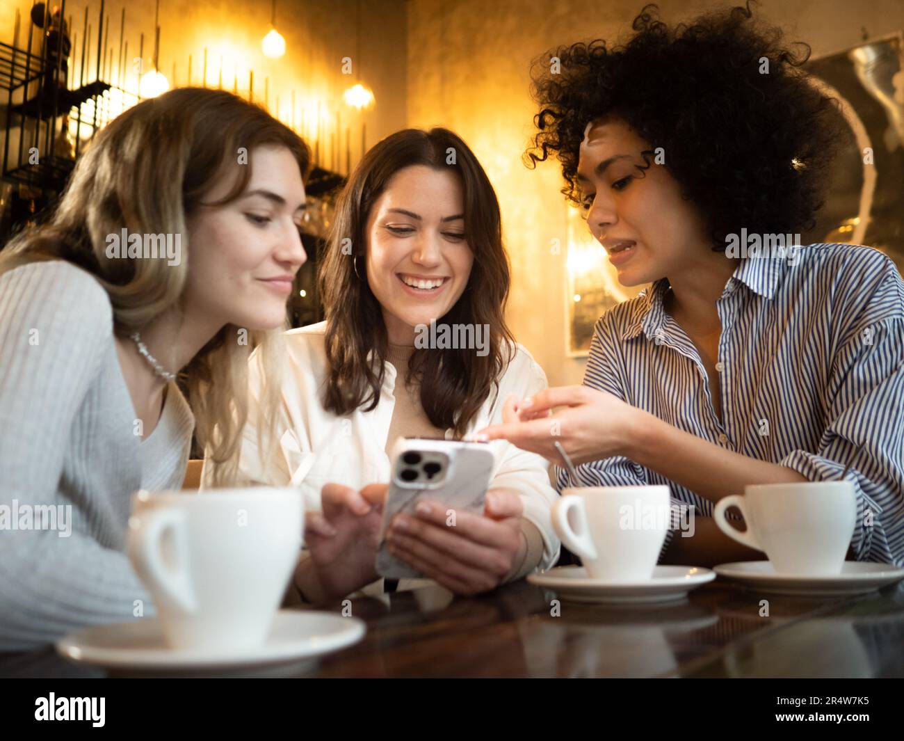 Trois jeunes femmes multiethniques se moque dans un café en train de rire et de parler tout en regardant le smartphone Banque D'Images