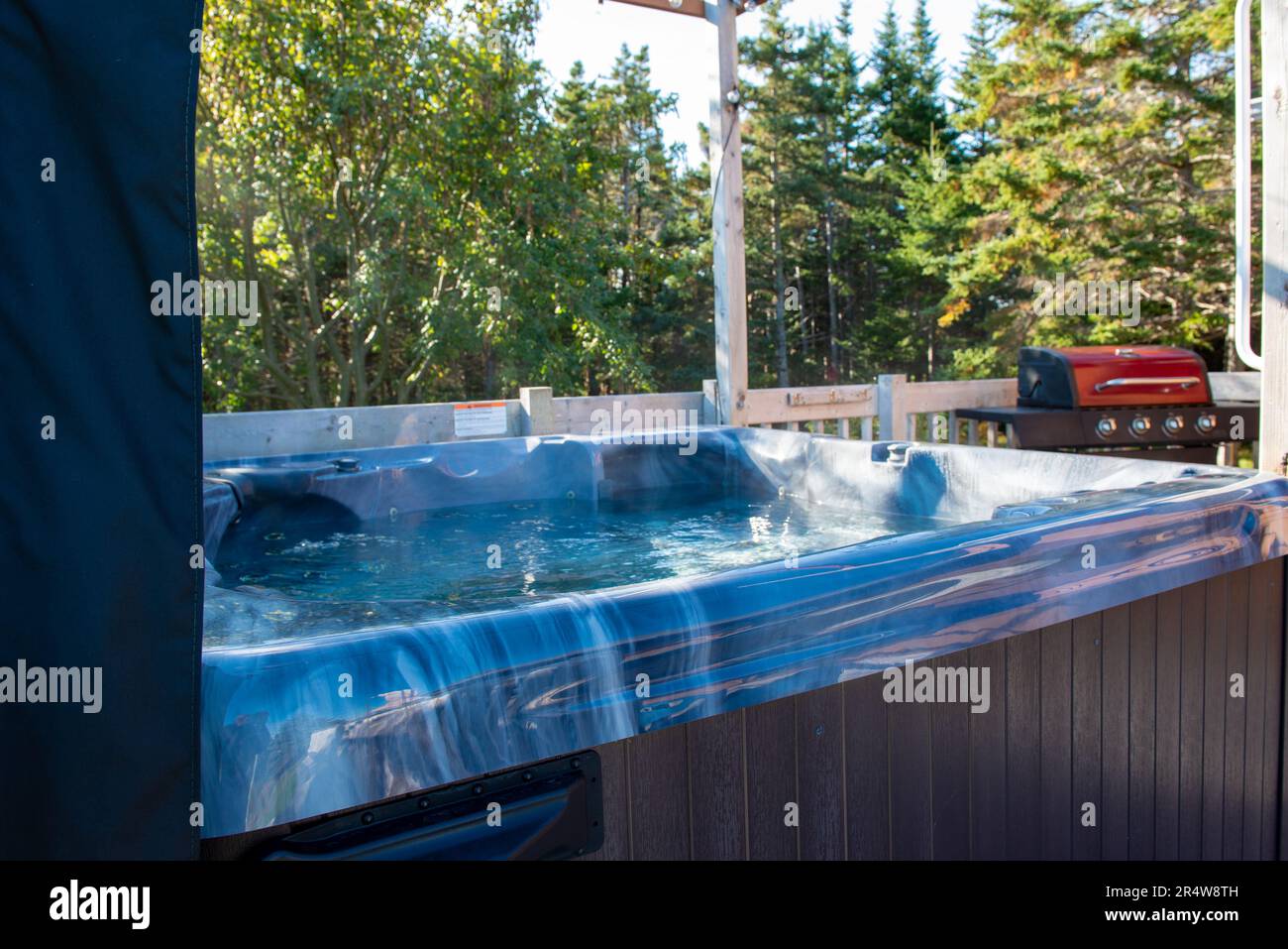 Un grand bain à remous bleu aqua vide sur une terrasse à côté d'un barbecue rouge. La vapeur de l'eau de la baignoire s'échappe de l'eau claire avec des bulles. Banque D'Images