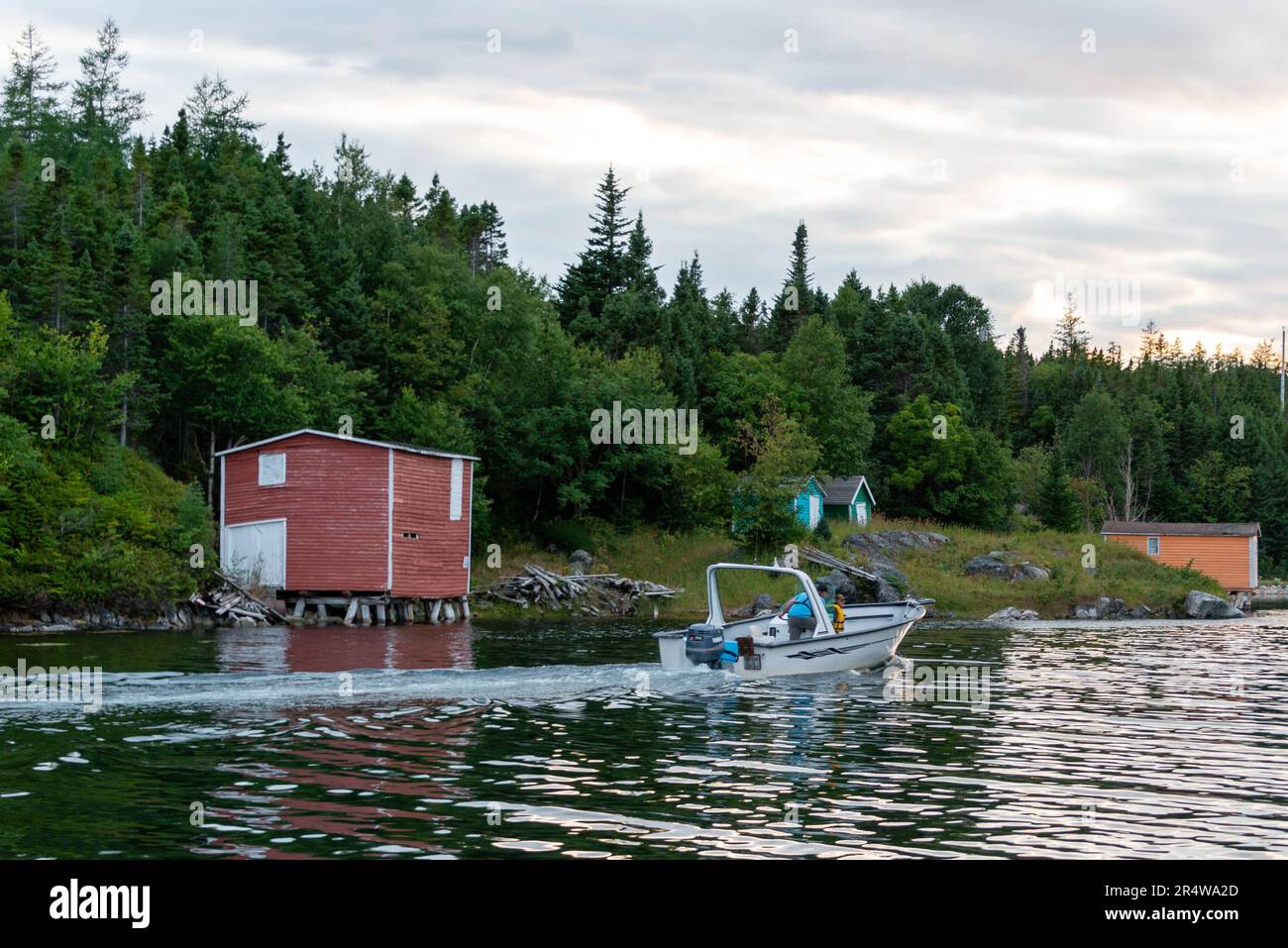 Un hangar à bateaux rouge avec une grande porte blanche sur le côté d'une rivière alors qu'un bateau à moteur remonte la rivière. La banque est couverte d'arbres verts luxuriants et d'un ciel gris. Banque D'Images