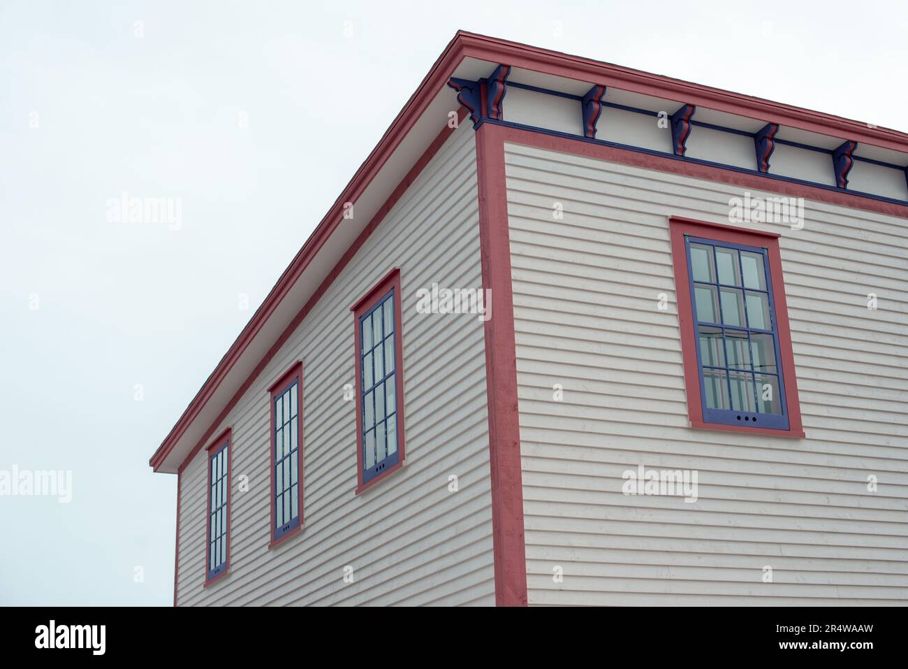 La section de toit d'un grand bâtiment blanc d'époque en bois avec des garnitures décoratives en bois rose et violet. Les fenêtres fermées sont en verre vintage à double pendaison. Banque D'Images