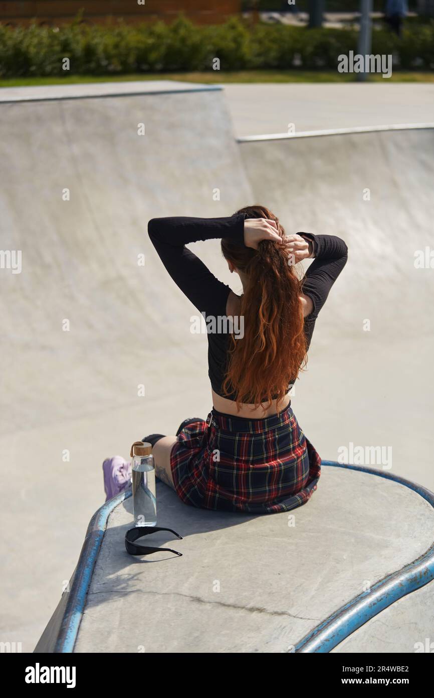 Patineuse femelle assise sur une rampe avec une bouteille d'eau en verre. Un jeune roller froid se rafraîchisseur dans un parc de skate par une belle journée d'été Banque D'Images