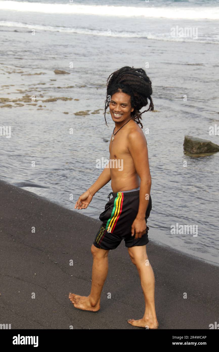 Asian Rasta Man marchant sur Bali Black Sand Beach en souriant à la caméra Banque D'Images