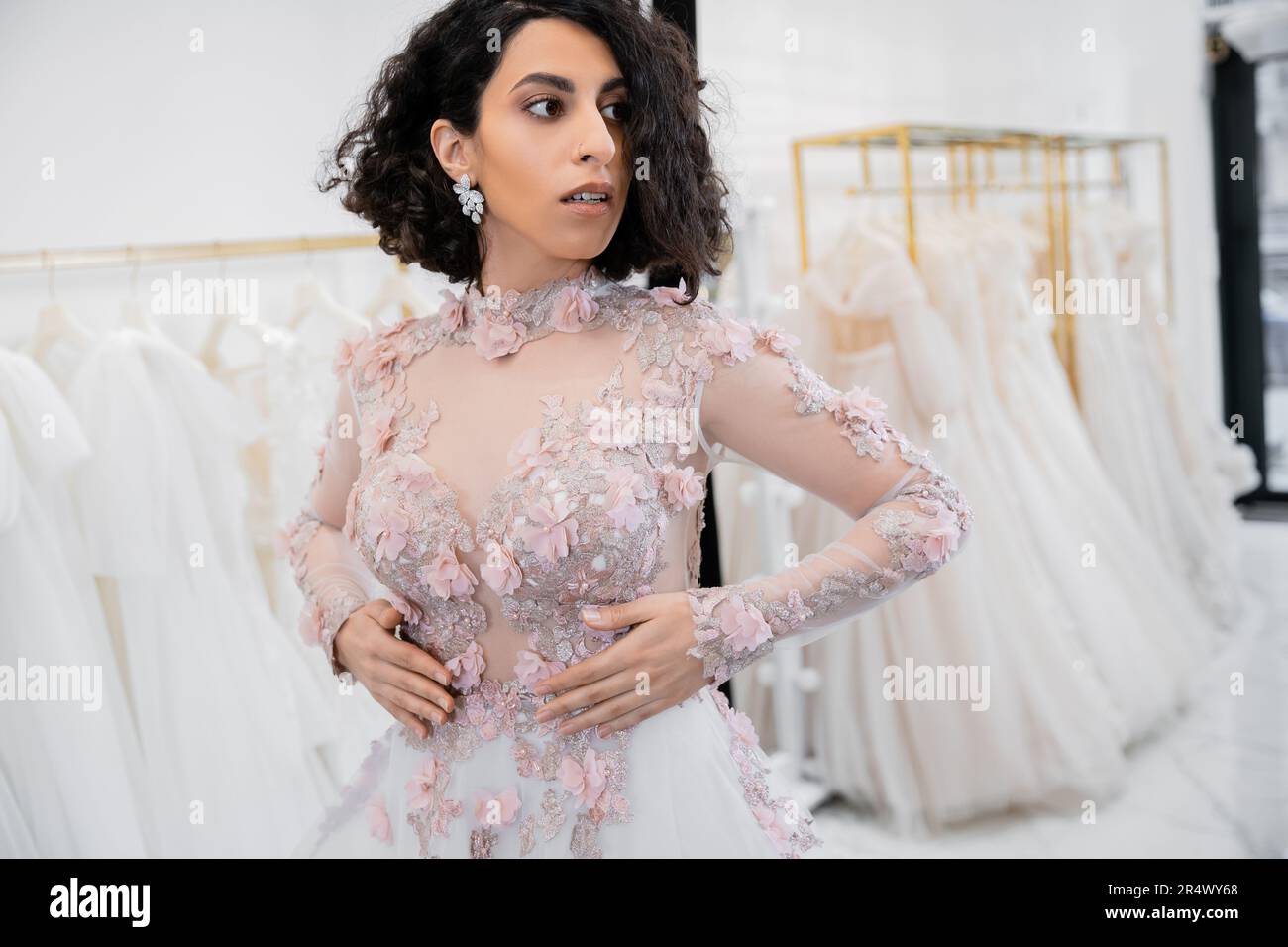 superbe mariée moyen-orientale et brune aux cheveux ondulés dans une magnifique tenue florale à l'intérieur d'un luxueux salon de mariage autour de la tulle blanche Banque D'Images