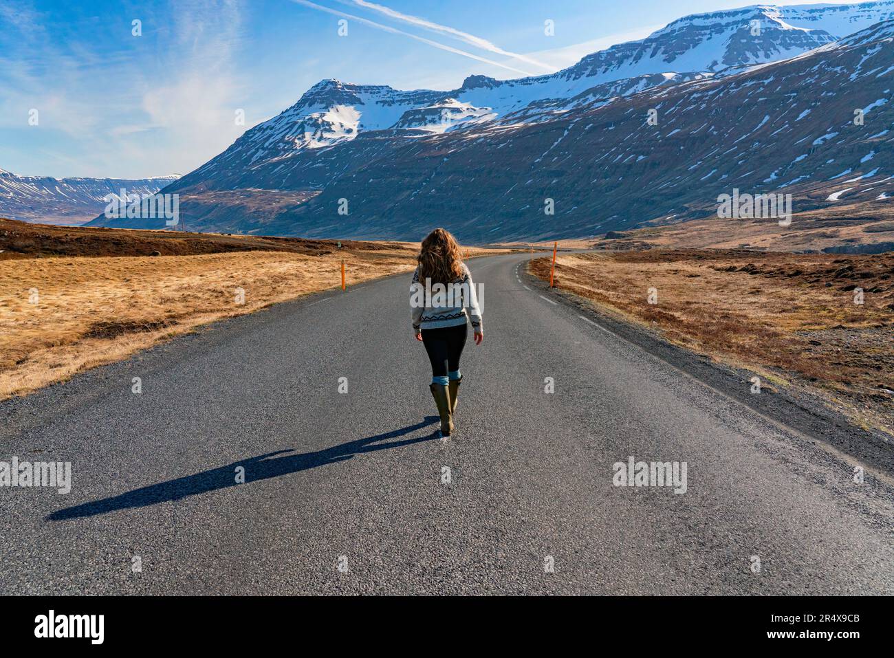 Femme marchant sur une route ouverte vers Seyðisfjörður avec une vue sur les montagnes enneigées et la beauté des fjords de l'est sous un ciel bleu Banque D'Images