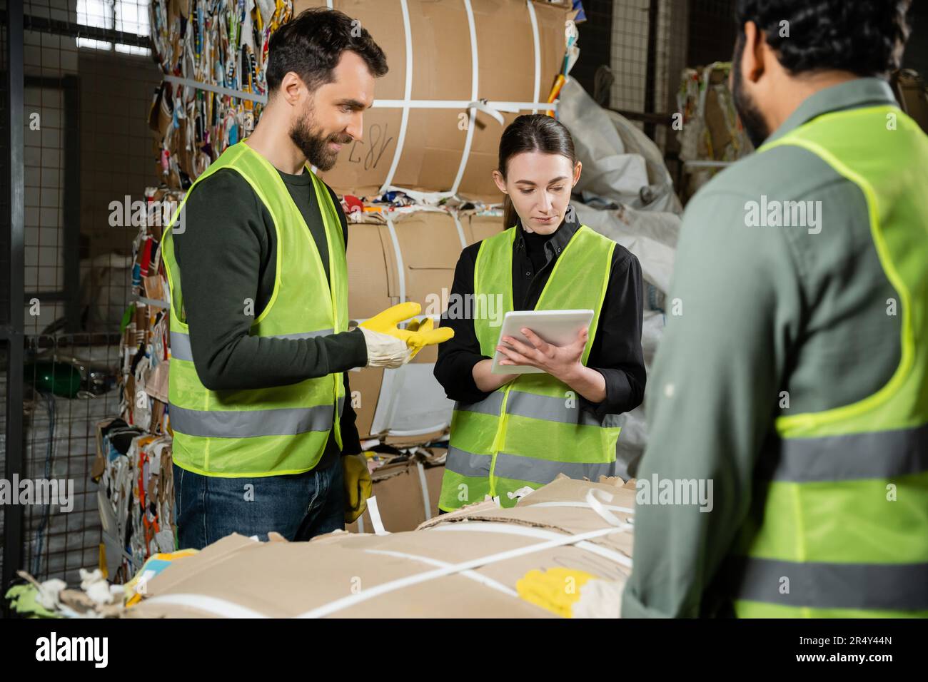 Jeune travailleur dans un gilet et des gants de protection utilisant une tablette numérique près de collègues multiethniques souriants et de déchets de papier dans le centre de tri des ordures, déchets Banque D'Images
