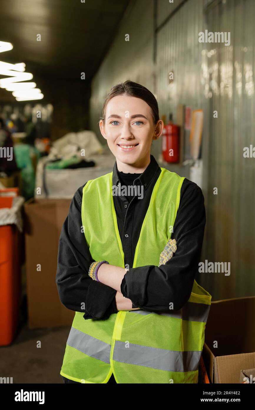 travailleur positif et jeune dans des gants et une veste haute visibilité croisant les bras et regardant la caméra tout en se tenant dans une station d'élimination des déchets floue à Banque D'Images