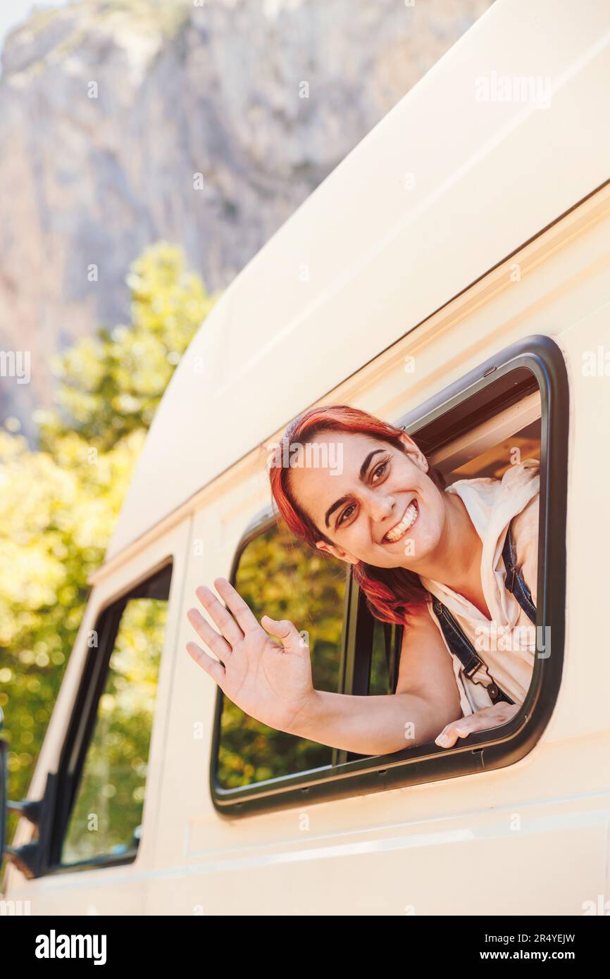 Une jeune femme agite par la fenêtre de sa camionnette de camping. Vacances en minibus et aventure en plein air en été. Concept de style de vie nomade Banque D'Images