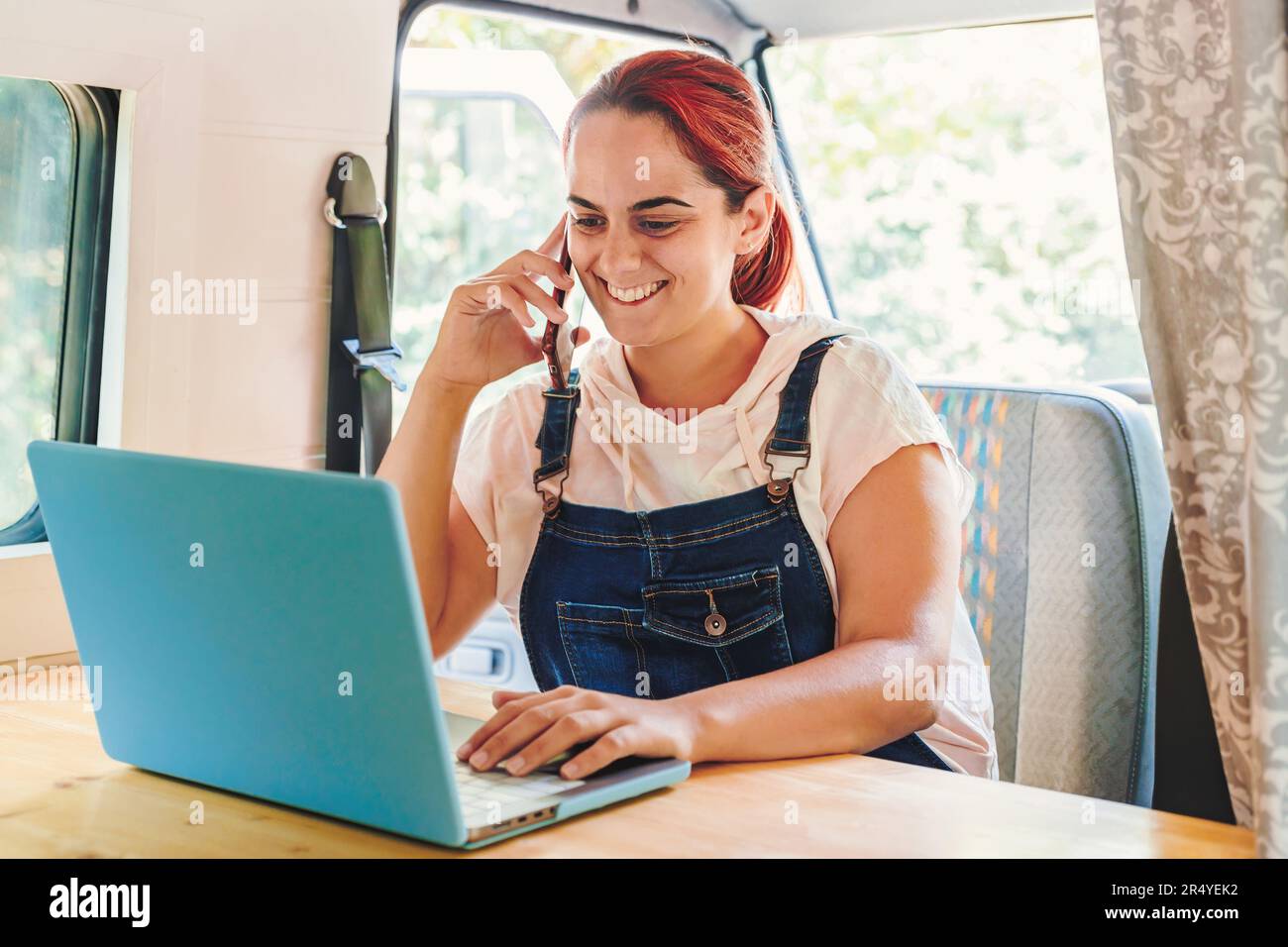 Femme travaillant sur son ordinateur portable et ayant un appel téléphonique dans sa camionnette de camping faite à la main pendant son voyage. Vacances en minibus et aventure en plein air en été. Non Banque D'Images
