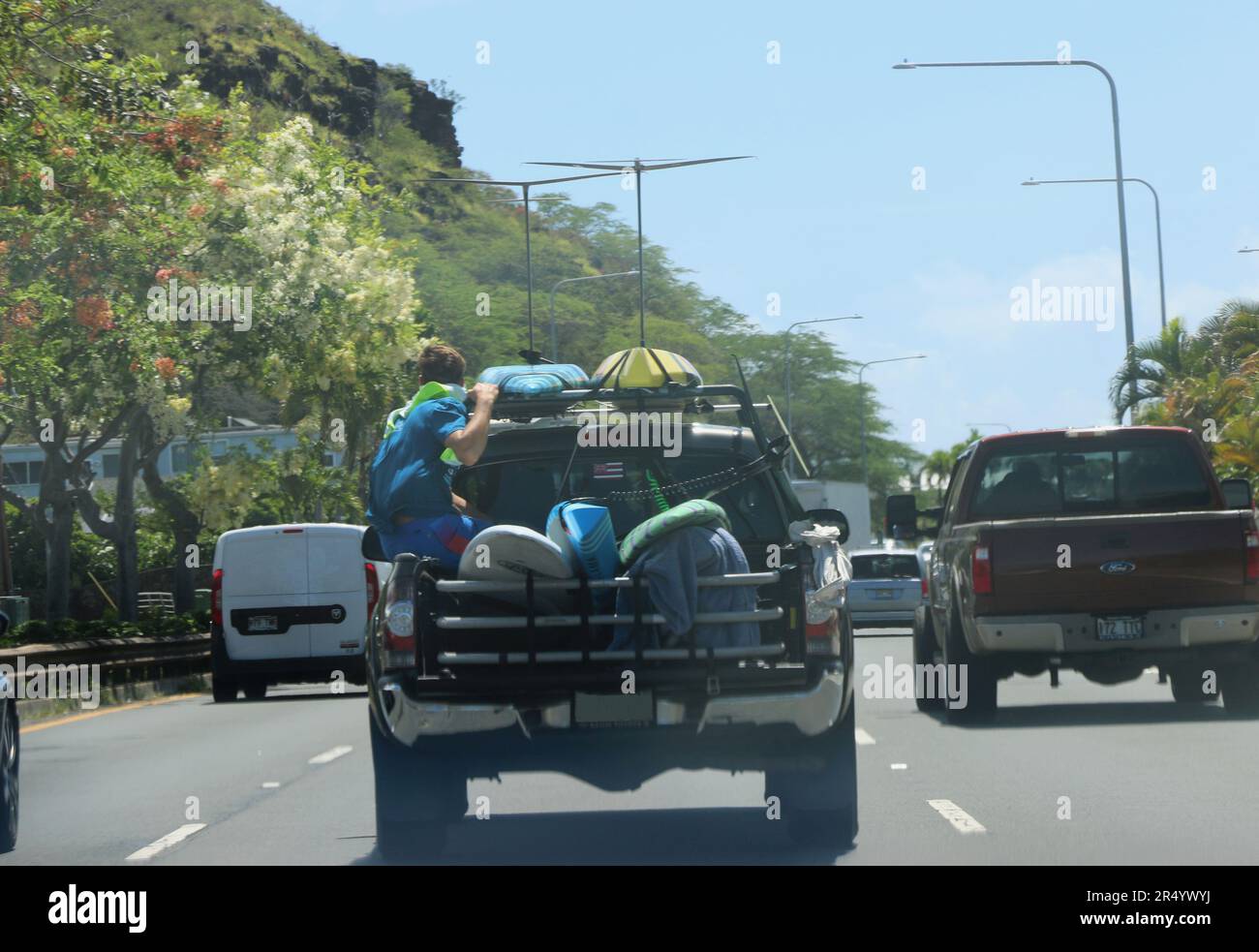 Honolulu, Hawaï, États-Unis - 30 mai 2023. Un jeune garçon se tient debout à l'arrière ouvert d'un camion accroché à des planches de surf sur une route très fréquentée. Banque D'Images