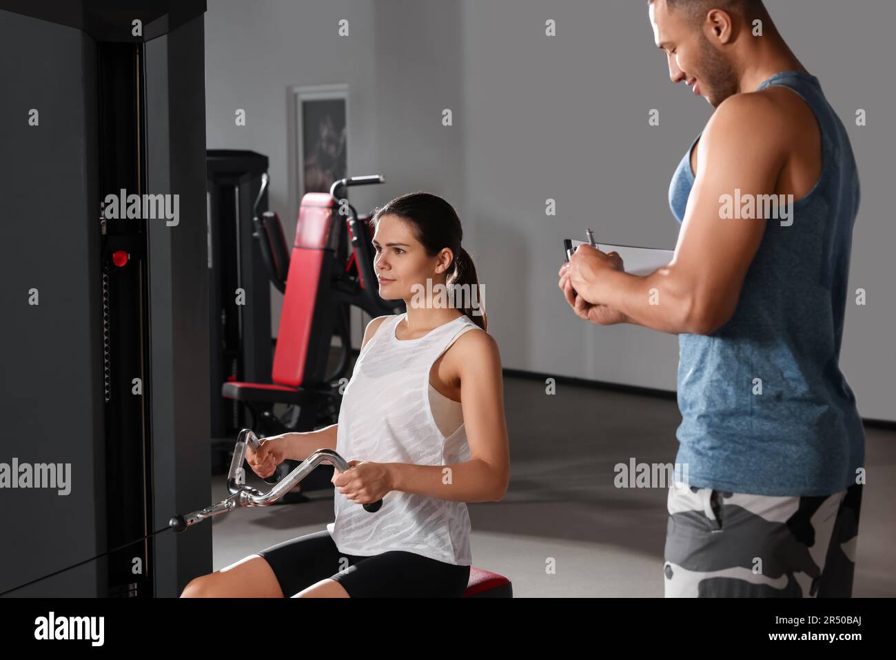 Entraîneur écrivant le plan d'entraînement pendant que la femme fait de l'exercice dans la salle de gym moderne Banque D'Images