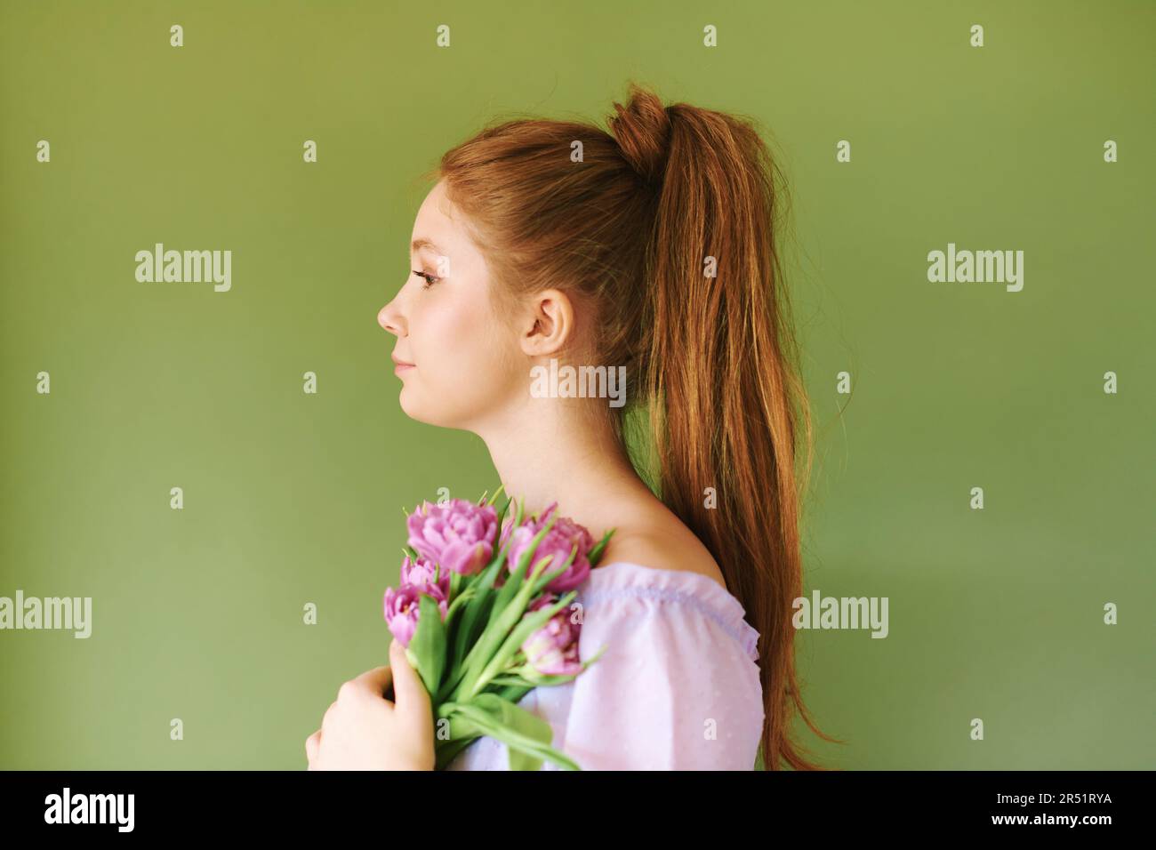 Portrait de beauté de la jolie jeune fille de 15 - 16 ans de téeenage à cheveux rouges vêtue de robe pourpre posant sur fond vert, tenant des fleurs de tulipe Banque D'Images
