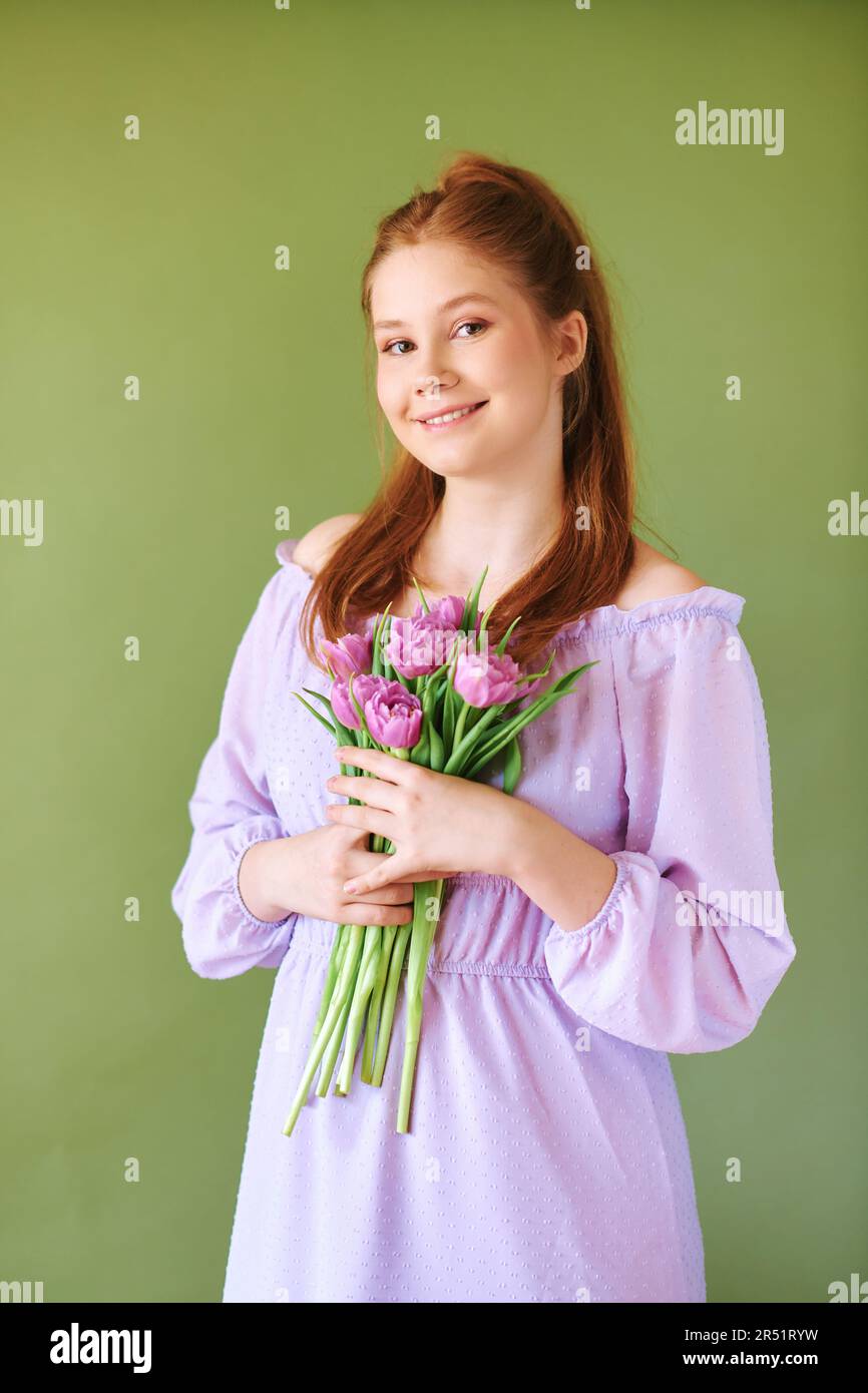 Portrait de beauté de la jolie jeune fille de 15 - 16 ans de téeenage à cheveux rouges vêtue de robe pourpre posant sur fond vert, tenant des fleurs de tulipe Banque D'Images