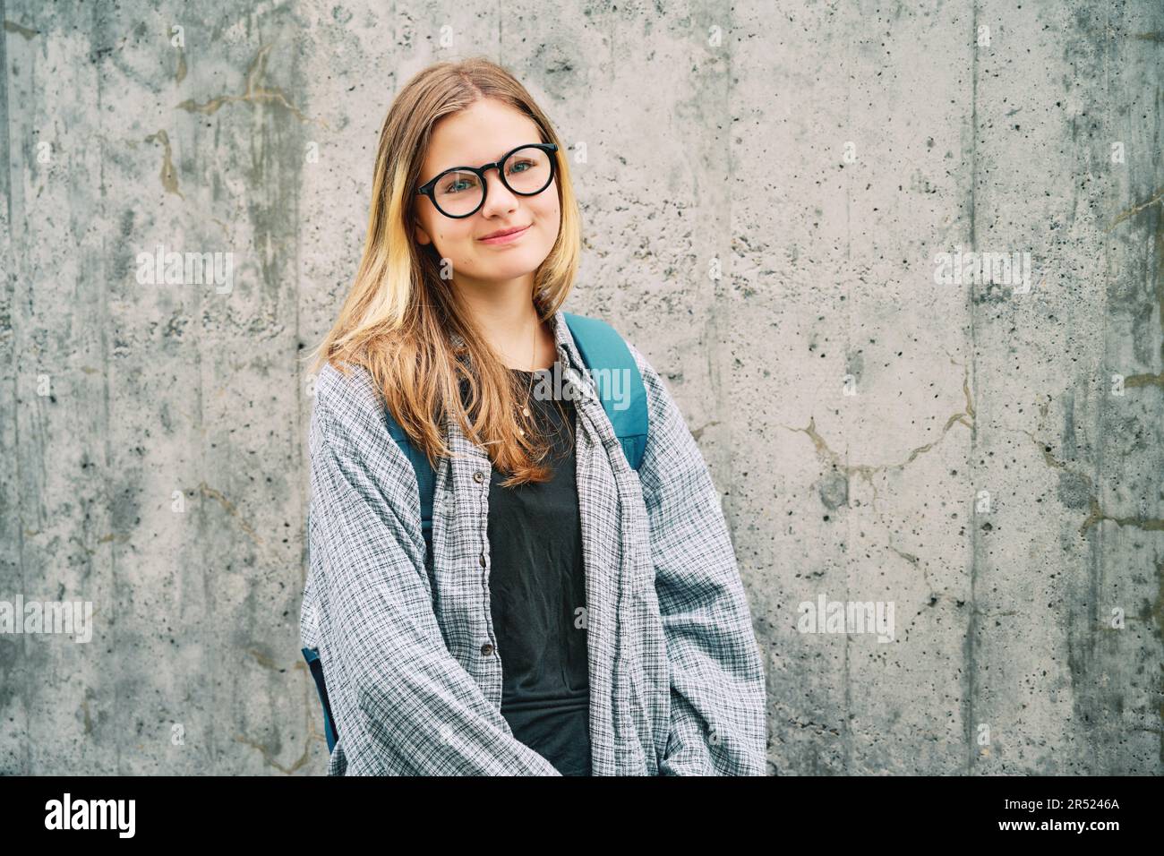 Portrait en plein air d'une jeune fille adolescente portant des lunettes et un sac à dos, posant sur un fond de mur gris Banque D'Images