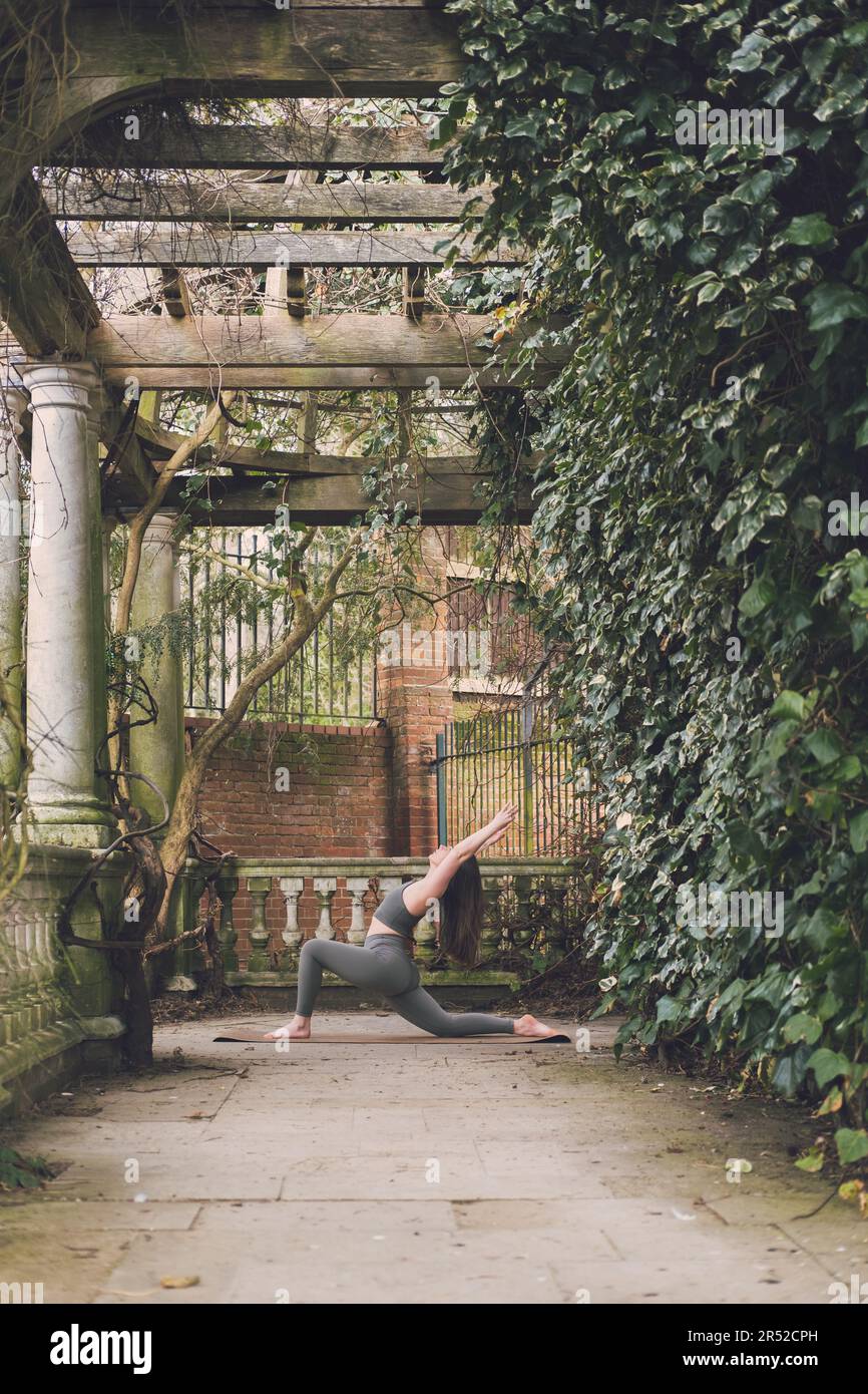 Enseignante de yoga en fente basse avec posture de flexion arrière pendant son entraînement en plein air seule avec tenue verte dans une belle terrasse d'un jardin paysagé Banque D'Images