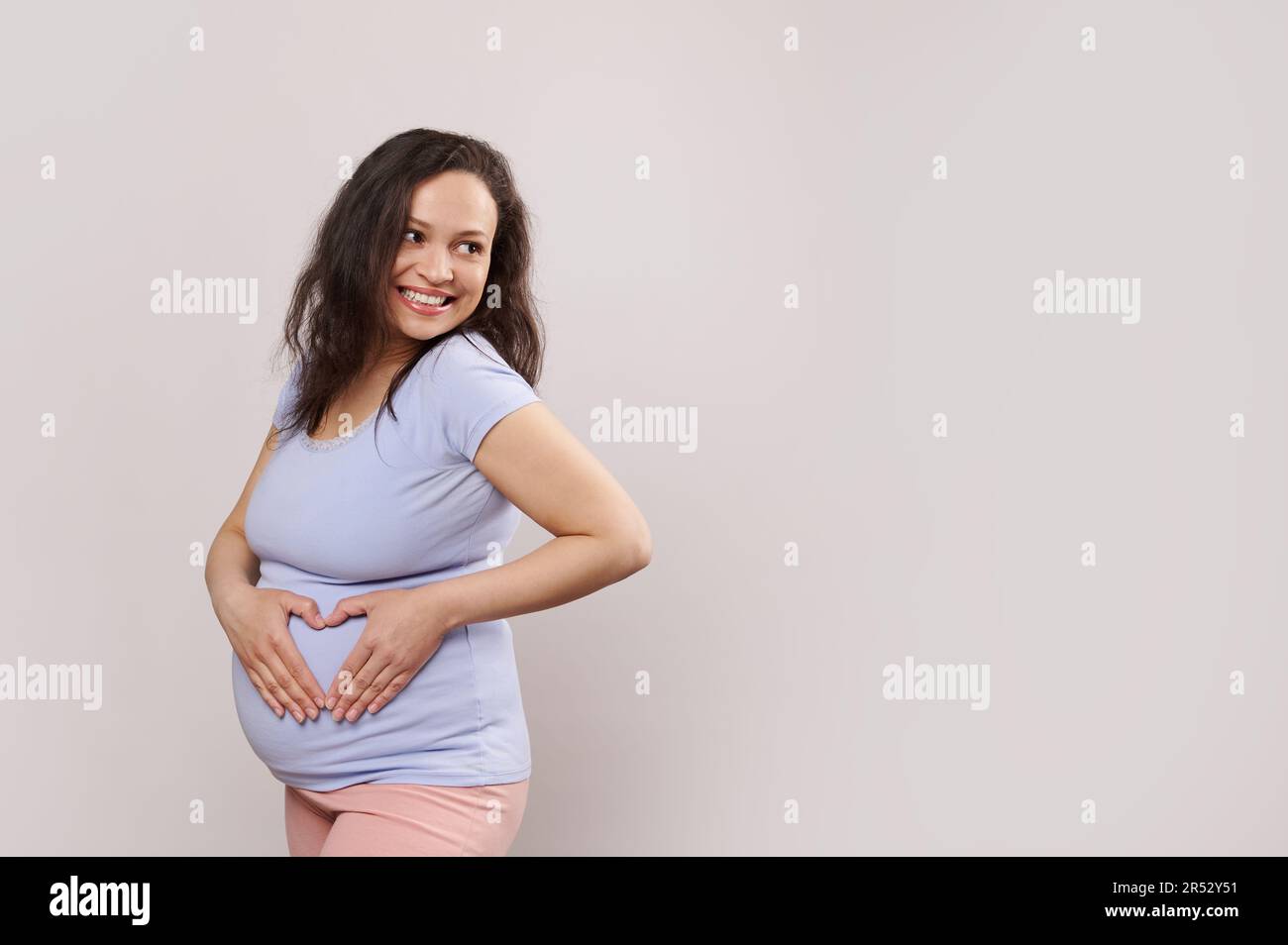 Bonne femme enceinte, femme enceinte souriante en regardant de côté, formant le coeur avec les mains sur le ventre, isolé sur blanc Banque D'Images
