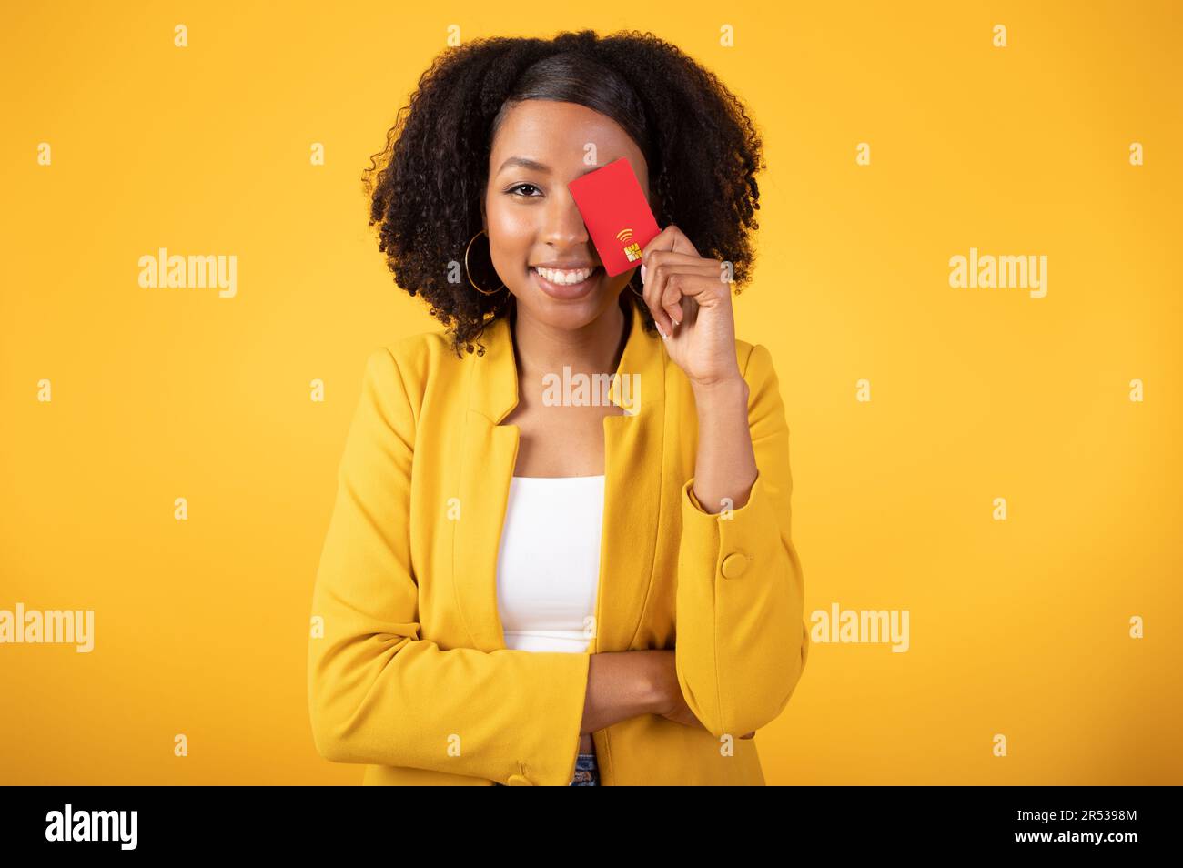 Paiements faciles. Jeune femme noire excitée couvrant l'œil avec carte de crédit et souriant à l'appareil photo, fond jaune Banque D'Images