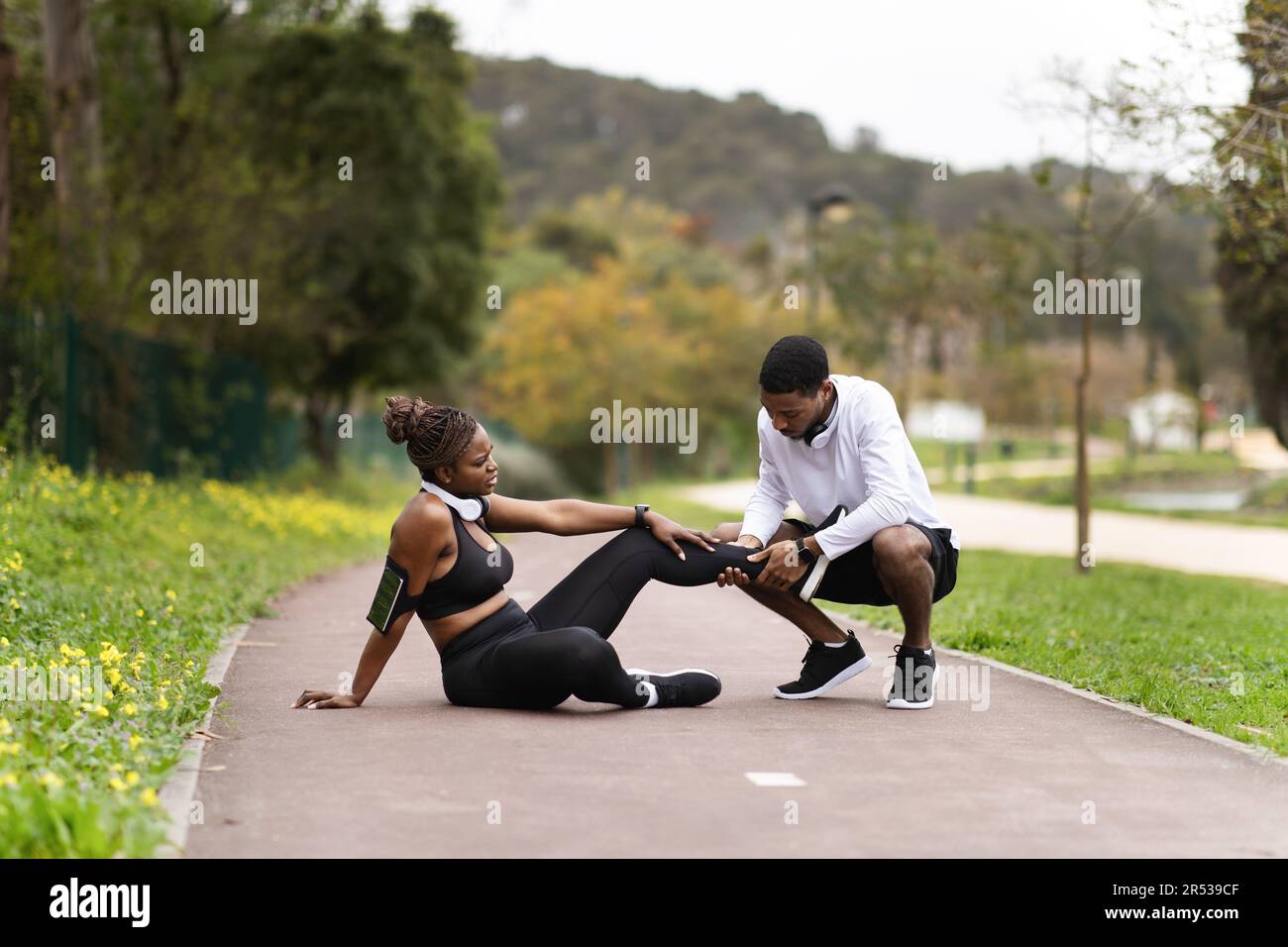 Accident et assistance. Jeune homme noir en vêtements de sport aide à la dame, souffrant de blessures aux jambes, faire un massage Banque D'Images