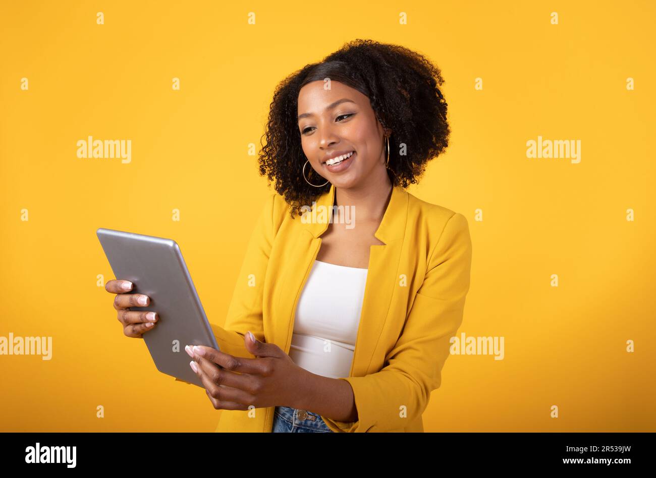 Joyeux étudiant noir tapant sur la tablette, regardant la vidéo et souriant isolé sur fond jaune, studio Banque D'Images