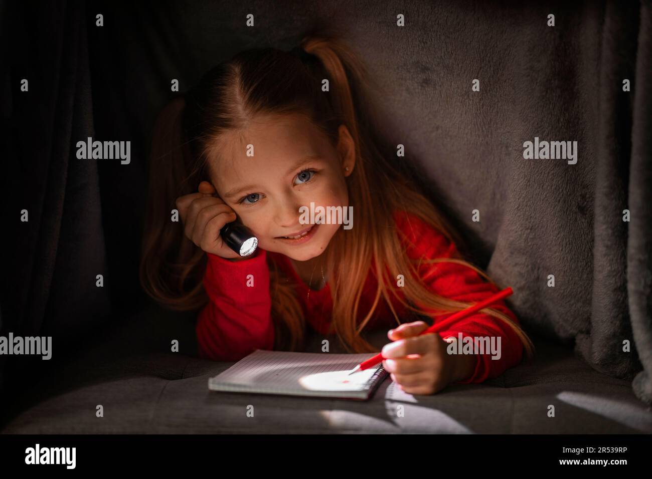 Jolie fille souriante masquage sous couverture avec lampe de poche et écriture dans le carnet Banque D'Images