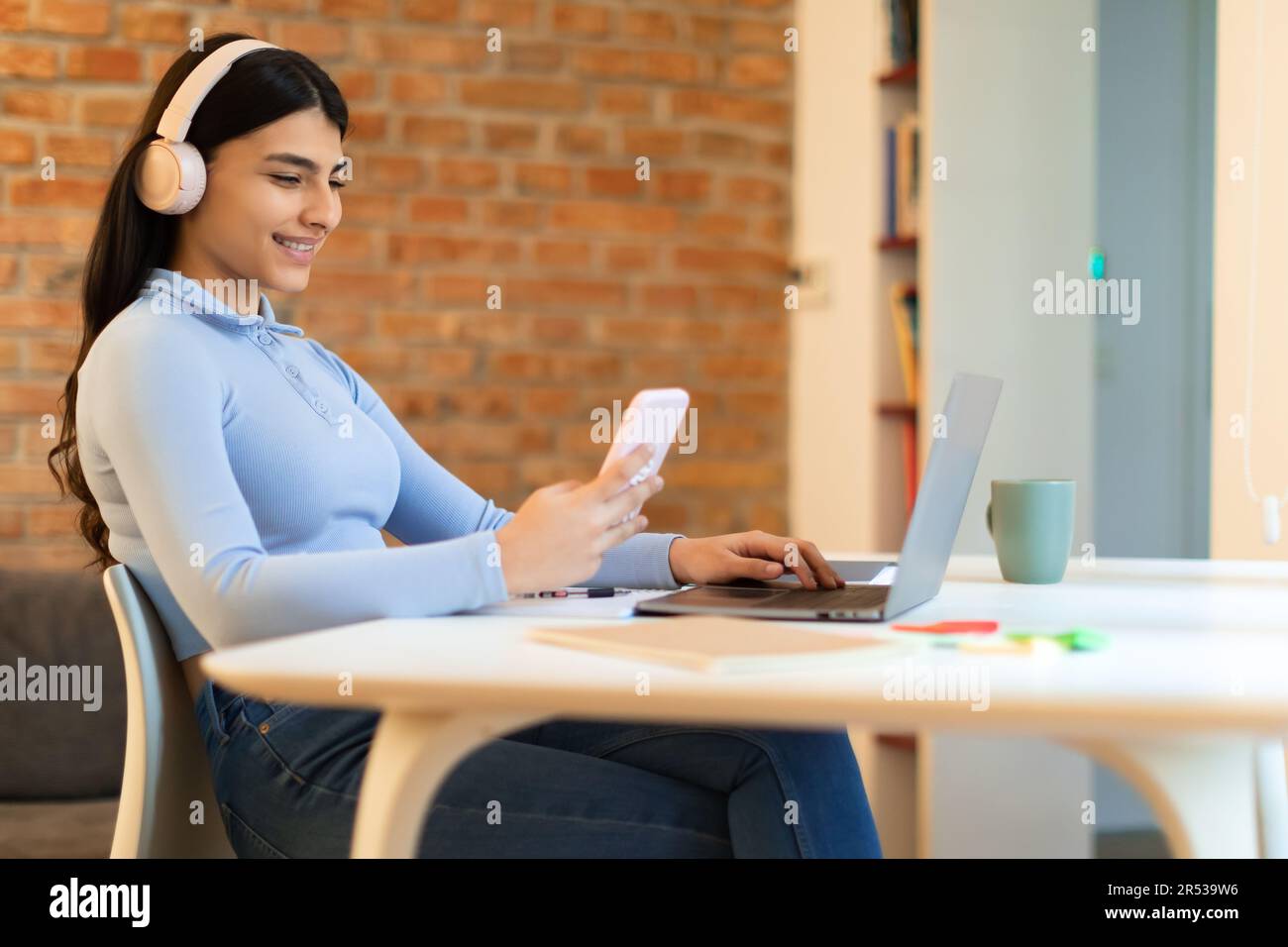Joyeuse adolescente espagnole étudiant de la maison, assis avec un ordinateur portable, casque et téléphone, ayant pause des cours Banque D'Images