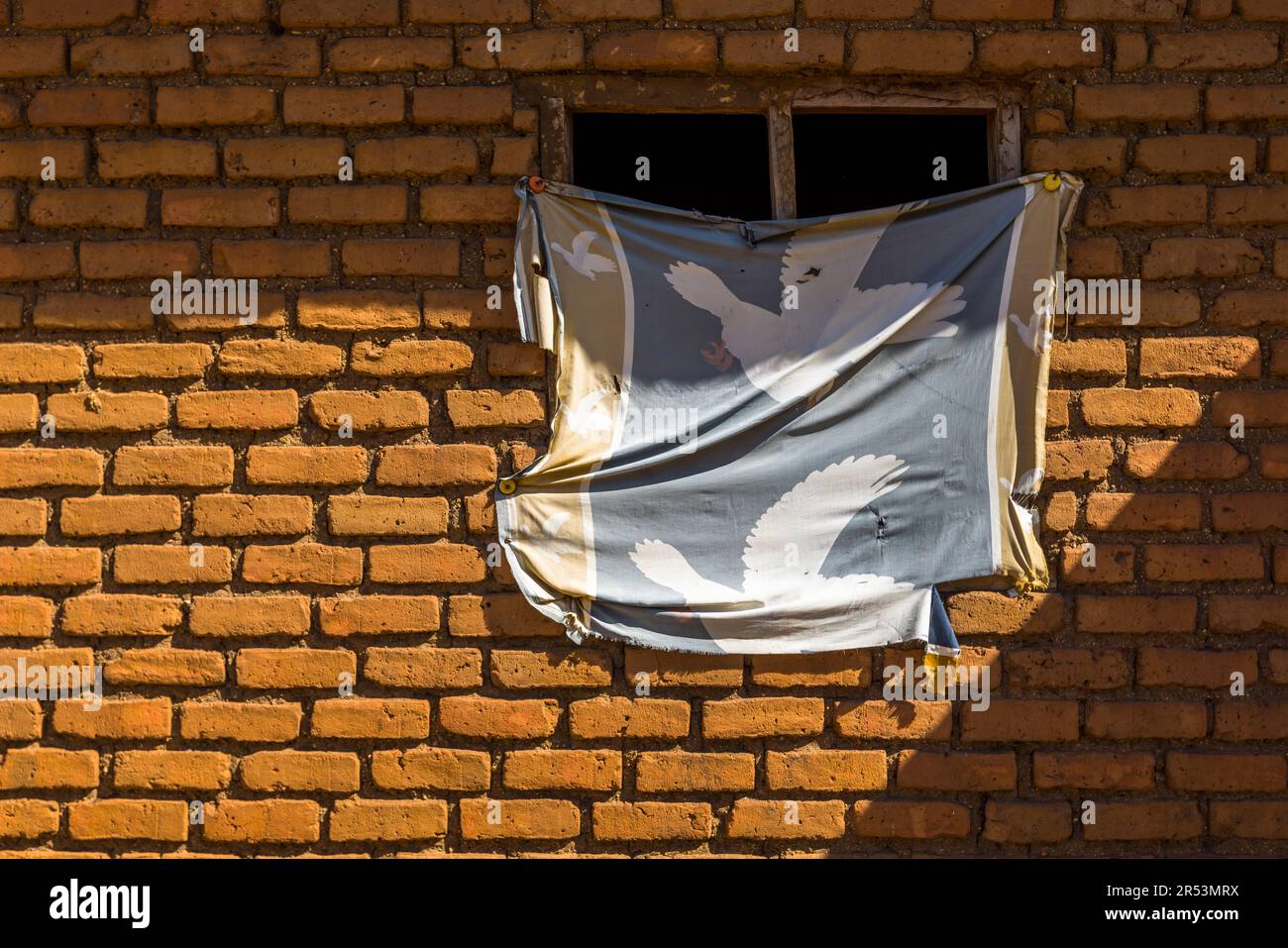 Un tissu imprimé d’un motif d’oiseau blanc est suspendu devant une fenêtre d’une maison en briques d’argile près de Monkey Bay, Malawi ; dans de nombreux villages, ces briques sont façonnées dans l’argile et cuites sur un feu de bois ouvert. Banque D'Images