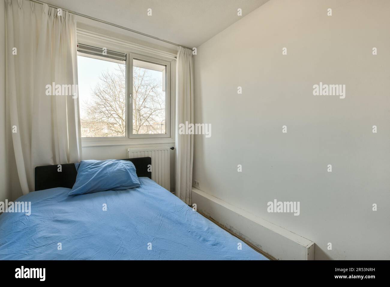un lit dans le coin d'une chambre avec des murs blancs et des draps bleus dessus, à côté d'une fenêtre Banque D'Images