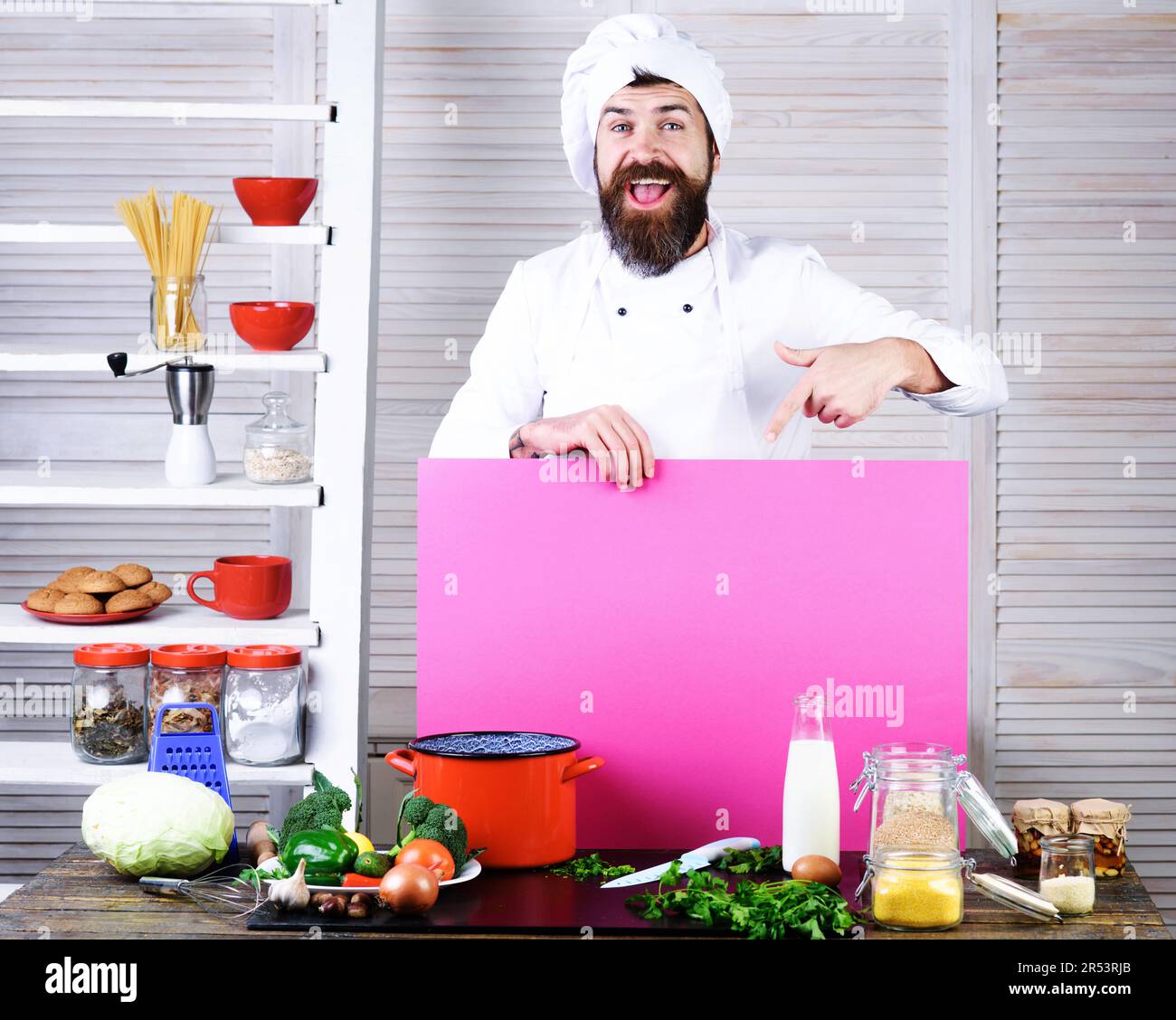 Un chef cuisinier à barbe heureux pointe le doigt vers un tableau vierge pour annoncer le menu. Chef en uniforme avec panneau d'affichage vide pour la publicité menu texte dans Banque D'Images