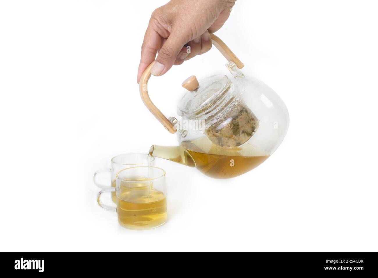 Quelqu'un main tenant la théière en verre puis versant le thé de fines herbes comme boisson saine à petit verre teacup sur fond blanc. Banque D'Images