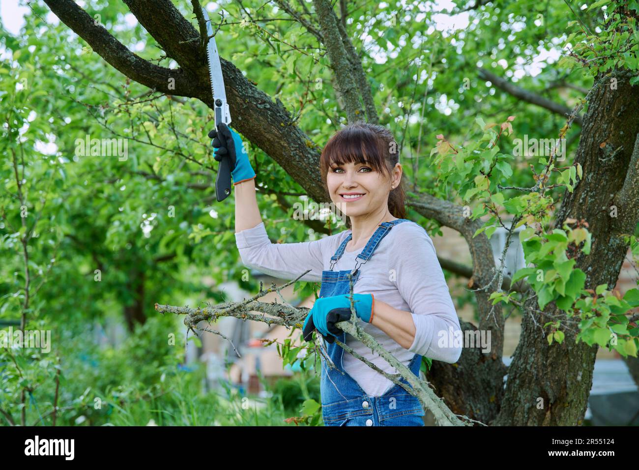 Femme jardinière avec une scie et des branches coupées à sec regardant la caméra dans le jardin Banque D'Images