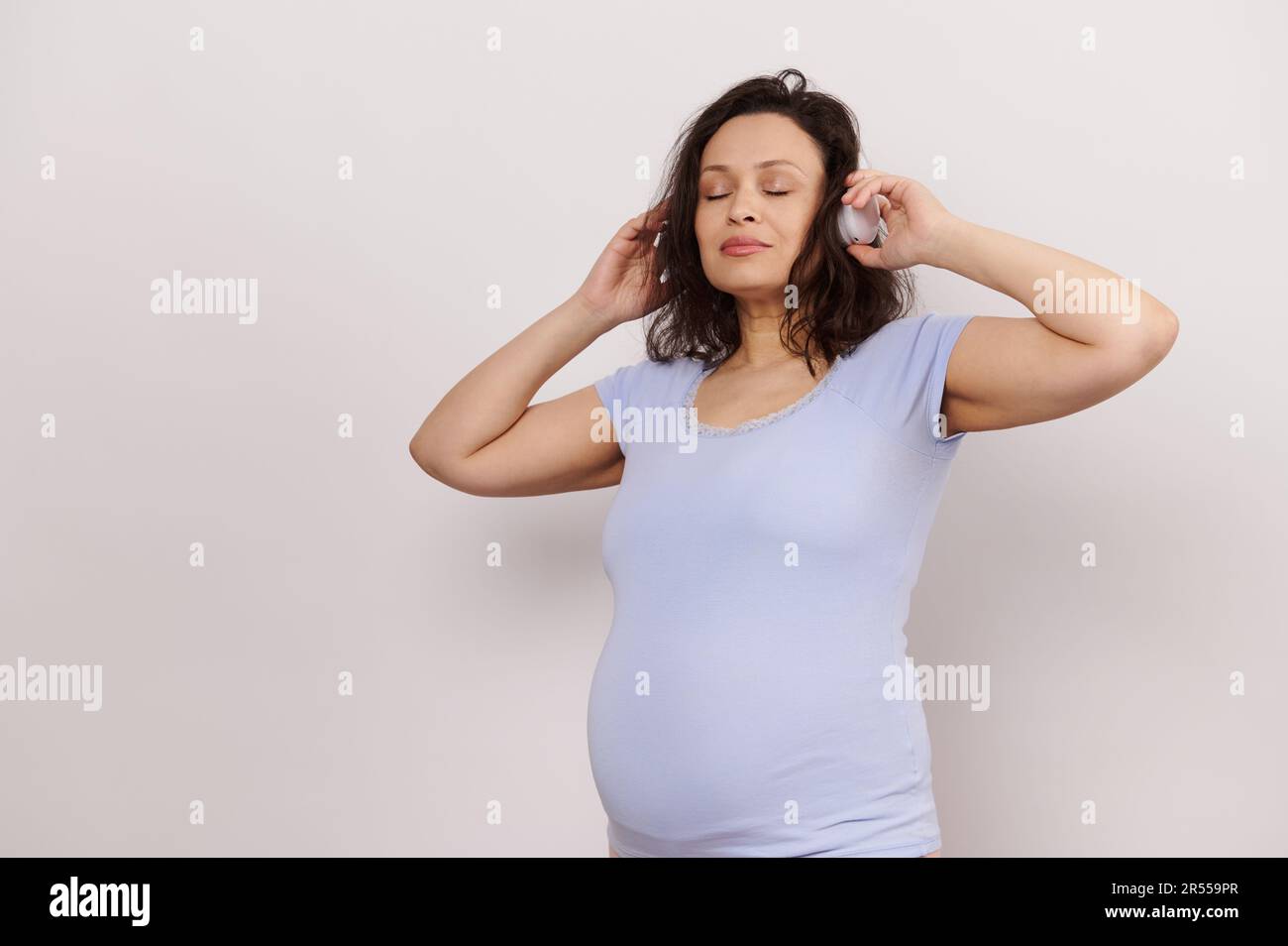 Bonne femme enceinte adulte qui écoute de la musique sur un casque sans fil, se posant avec ses yeux fermés sur fond blanc Banque D'Images