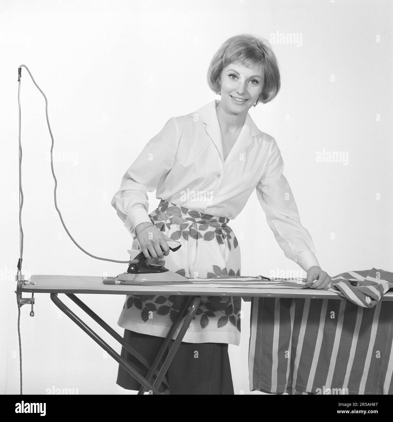 Repassage dans le 1960s. Une femme a vu des rideaux de repassage, souriant à l'appareil photo dans le studio de photographes. Suède 1965. Conard réf. 1743 Banque D'Images