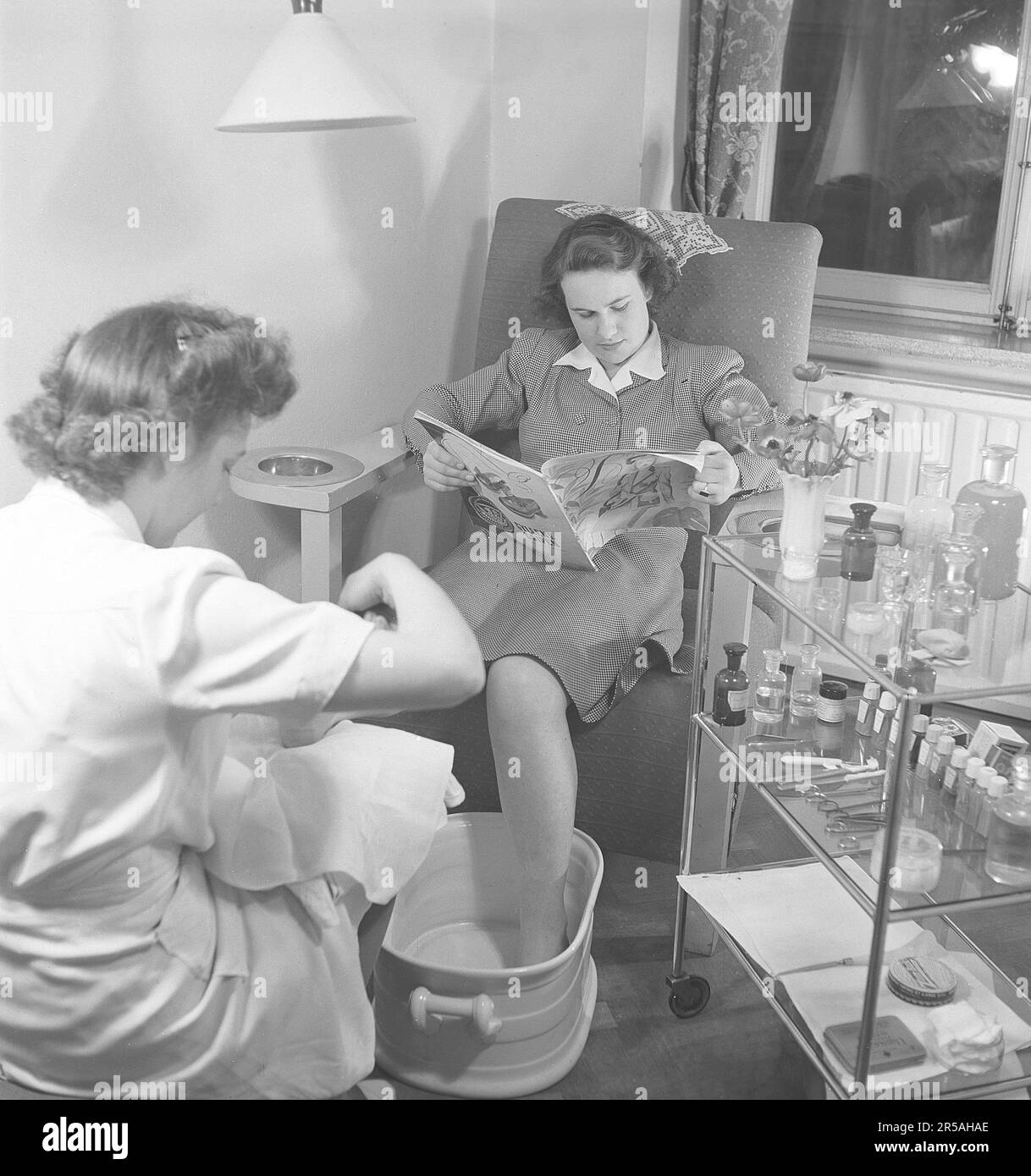 Une jeune femme dans un salon de beauté reçoit une pédicure. Le jeune employé effectue un traitement de soins des pieds où la tâche est d'embellir les pieds et les orteils. Une pédicure aide à garder vos pieds en bonne forme et les rend plus attrayants. Pendant que la jeune femme reçoit le traitement, elle s'assoit confortablement dans un fauteuil et lit un magazine. Suède 1945. Kristoffersson réf. N108-4 Banque D'Images