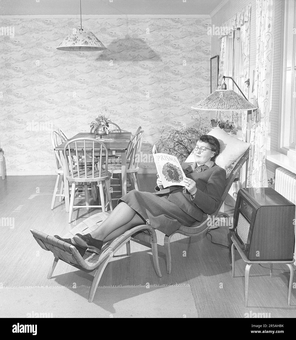 Décoration ancienne. Une salle de séjour où une femme se trouve dans la chaise Pernilla classique de Bruno Mathsson avec repose-pieds et lit un journal. Une radio se trouve sur une table à côté. Les lampes ont des abat-jour en tissu plissé et dans la même pièce aussi une table à manger avec des chaises assorties. Suède 1950. Kristoffersson réf. AX79-3 Banque D'Images