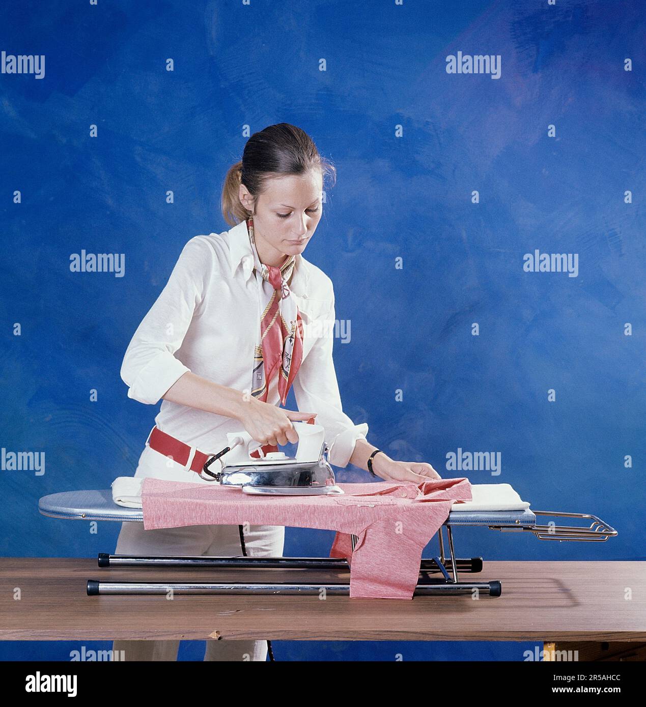 Repassage dans le 1970s. Une femme a vu repasser ses vêtements. Elle est habillée d'une chemise et d'un pantalon blancs et d'une ceinture rouge avec des pinces en métal, un accessoire typique de la décennie. Suède 1971. Kristoffersson réf. BV79-5 Banque D'Images