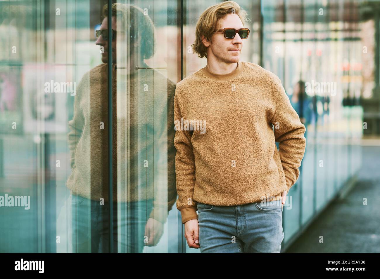 Portrait extérieur d'un jeune homme élégant portant des lunettes de soleil et un chandail en polaire beige, posé à côté du mur miroir sur le fond de la rue de la ville Banque D'Images