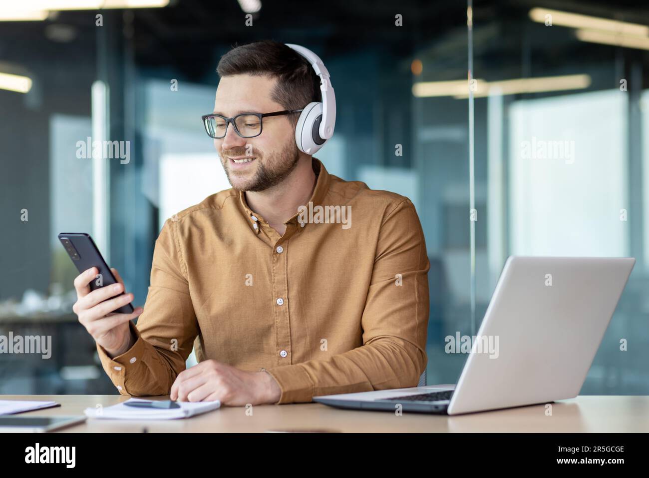 Un jeune homme souriant dans un casque et avec un téléphone dans les mains était assis à un bureau dans le bureau. Il travaille, se repose, parle sur un appel vidéo, écoute de la musique. Banque D'Images