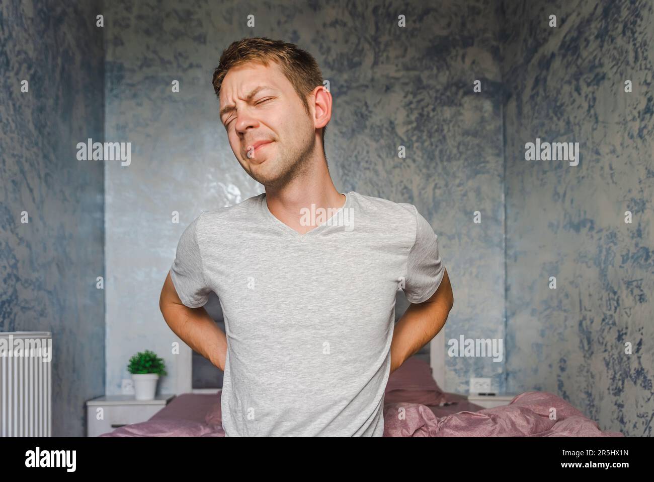 Jeune homme caucasien souffrant de maux de dos tout en étant assis sur un lit à la maison. Douleur de rein, mal de dos, lumbago concept Banque D'Images