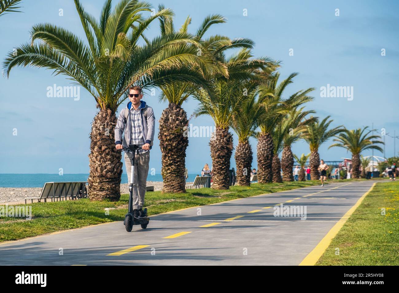 Jeune homme voyageur à cheval électrique le long de la côte de mer et de la plage à la journée ensoleillée d'été. Homme conduisant e- scooter à l'extérieur. Vert durable Banque D'Images