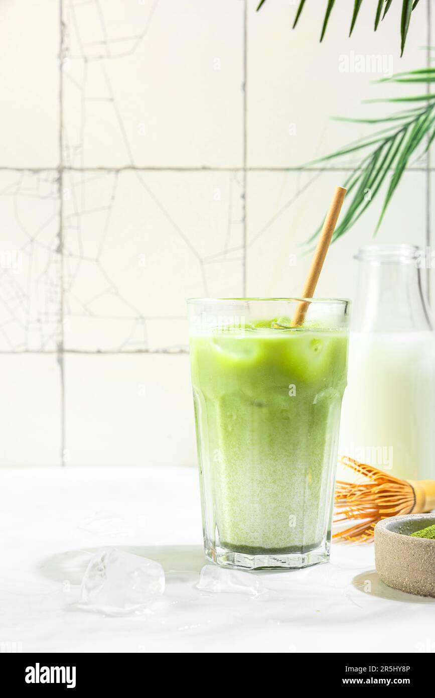 Latte matcha vert glacé dans un verre sur fond de carreaux blancs avec des ombres dures et des feuilles de palmier. Boisson d'été froide Banque D'Images