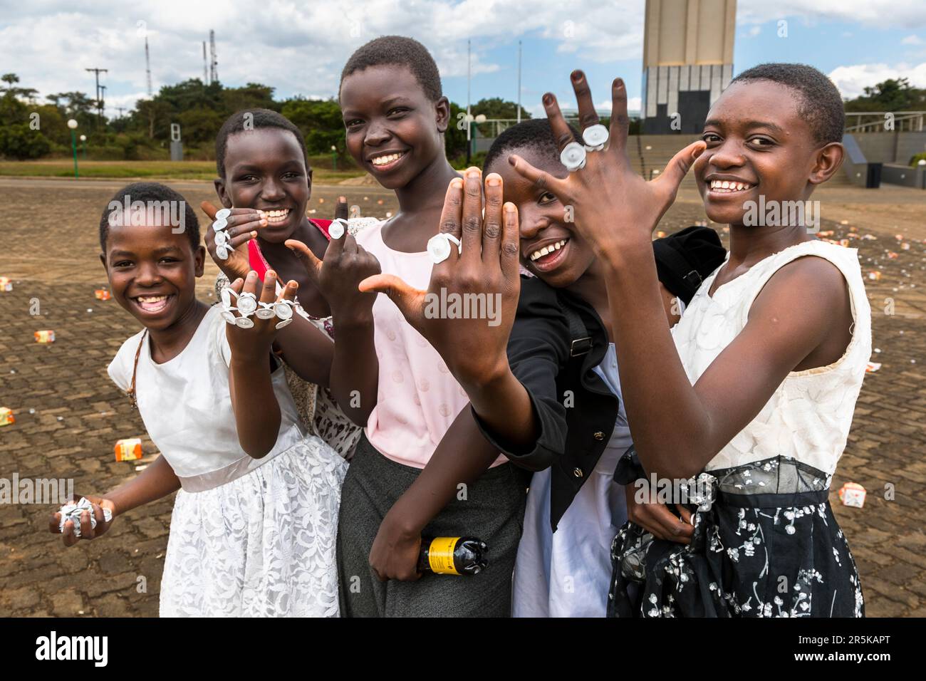 Cinq filles tendent leurs mains ornées de bagues en plastique faites maison à partir de capsules de bouteilles de Maheu, riant vers l'appareil photo devant la Tour du Mémorial National de la Guerre à Lilongwe, au Malawi. Maheu est une boisson traditionnelle malawienne faite à partir de maïs fermenté. Banque D'Images