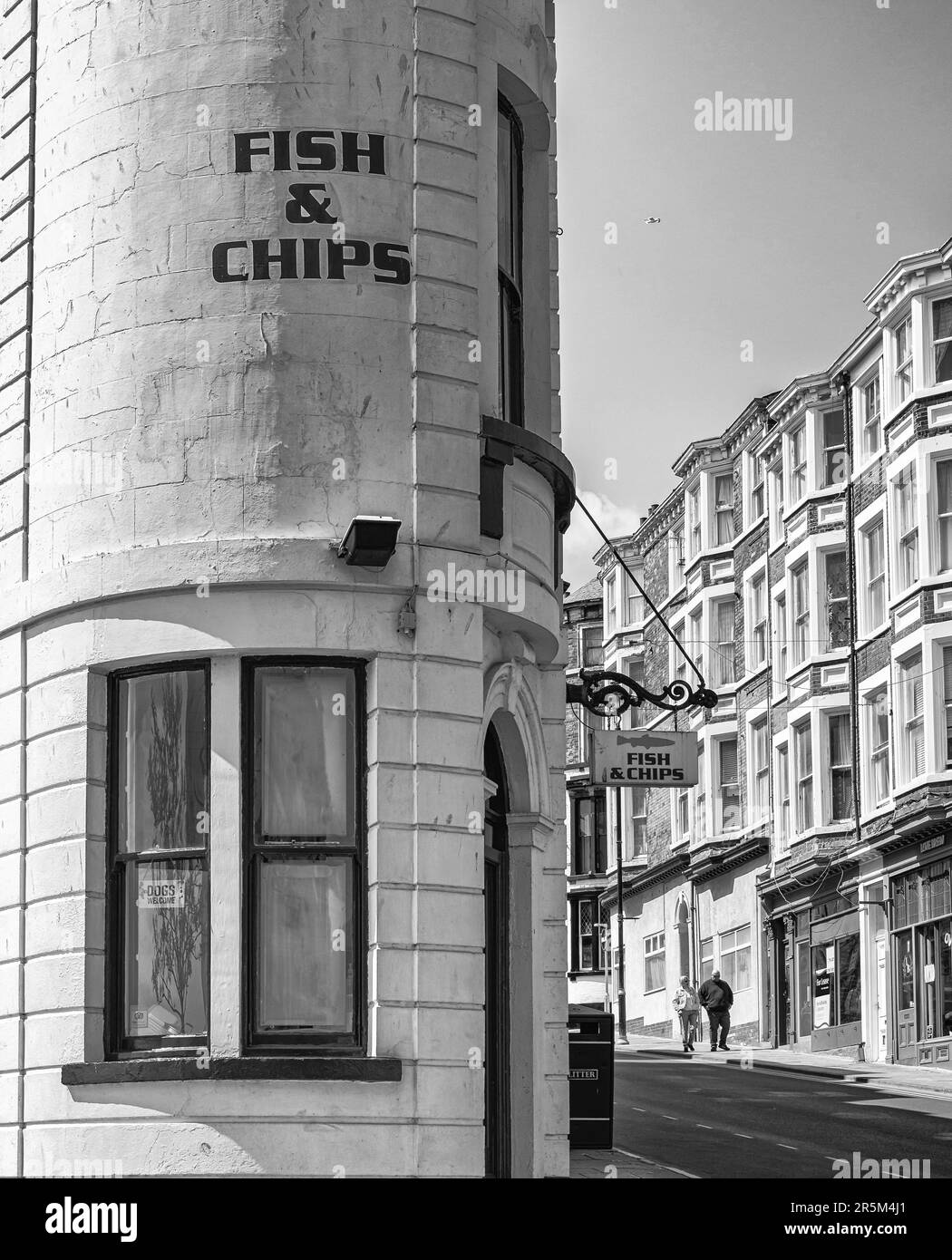 Un restaurant de poissons et de chips dans un bâtiment incurvé sur une rue en pente. Les bâtiments en brique rouge sont en face avec des magasins et un couple de marche en bas de la chaussée. Banque D'Images