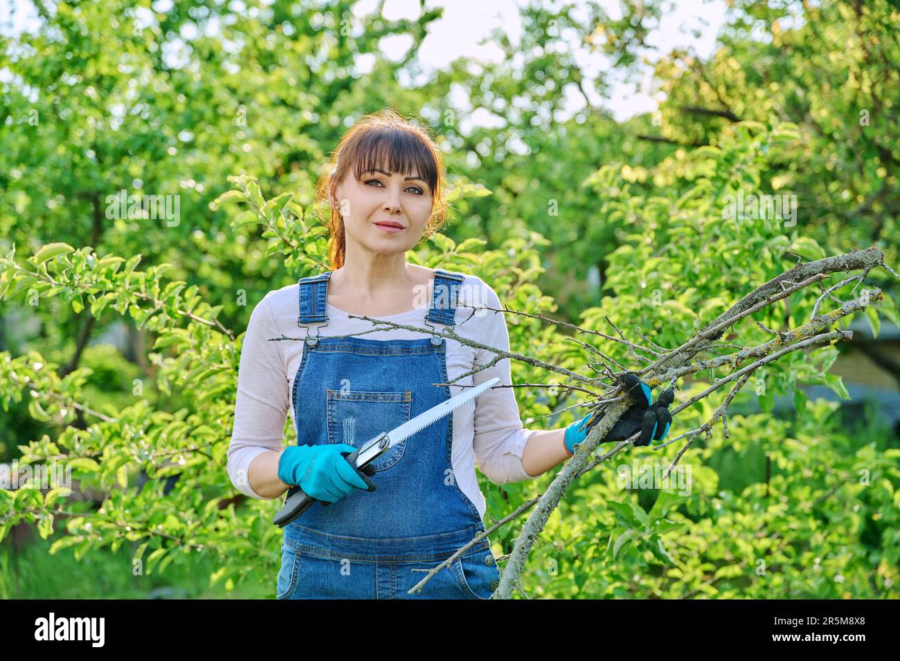 Femme jardinière avec une scie et des branches coupées à sec regardant la caméra dans le jardin Banque D'Images