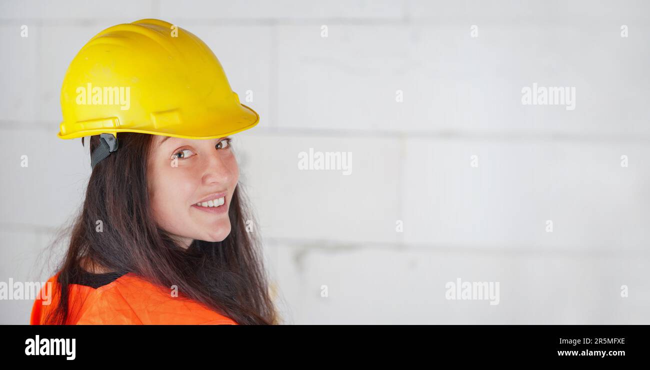 Jeune femme en casquette jaune rigide et gilet orange haute visibilité, cheveux longs foncés, regardant au-dessus de son épaule, souriant confiant. Construction floue assis Banque D'Images