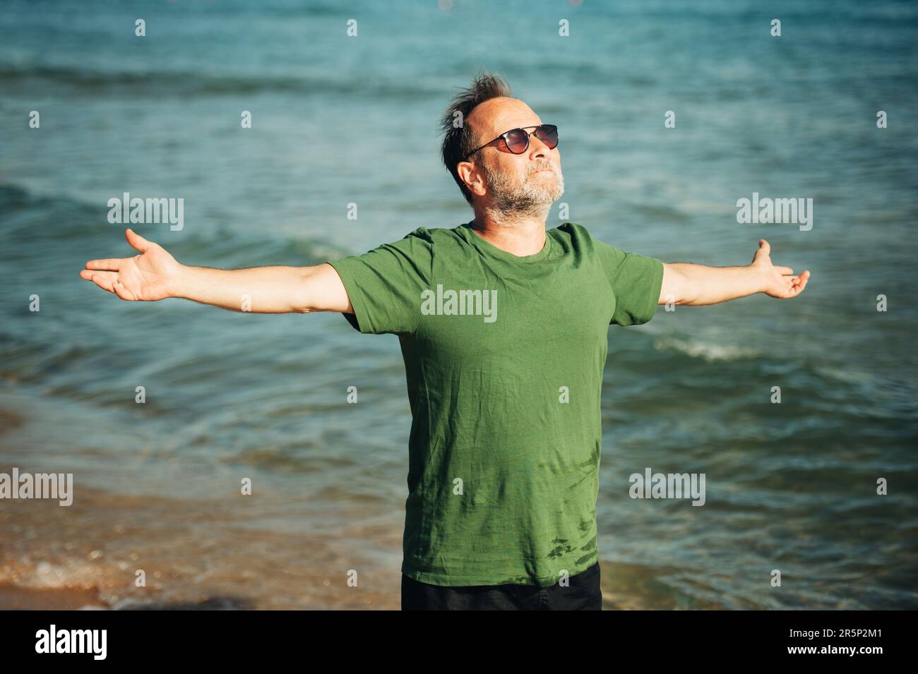 Portrait d'un homme heureux d'âge moyen bénéficiant d'une belle journée ensoleillée sur la plage, portant un t-shirt vert et des lunettes de soleil, les bras larges ouverts Banque D'Images