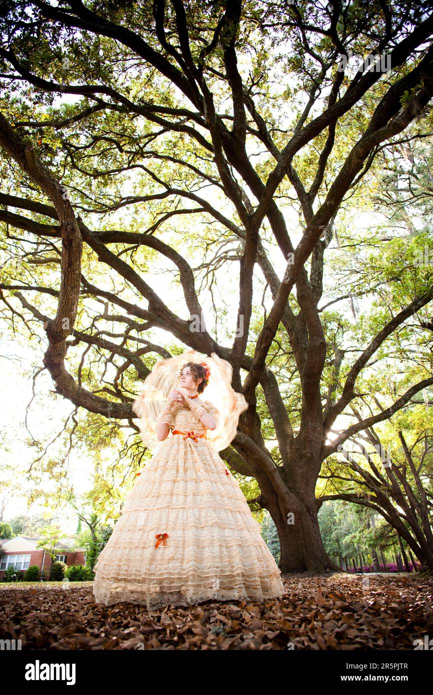 Une jeune femme pose dans une tenue Belle Sud , comprenant une grande robe d'époque ornée et un parasol, sous un arbre. Banque D'Images