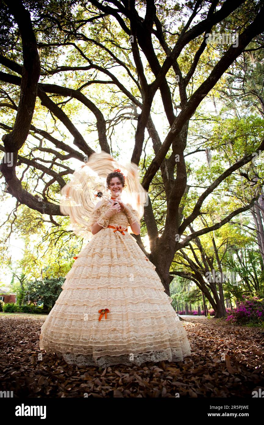 Une jeune femme pose dans une tenue Belle Sud , comprenant une grande robe d'époque ornée et un parasol, sous un arbre. Banque D'Images