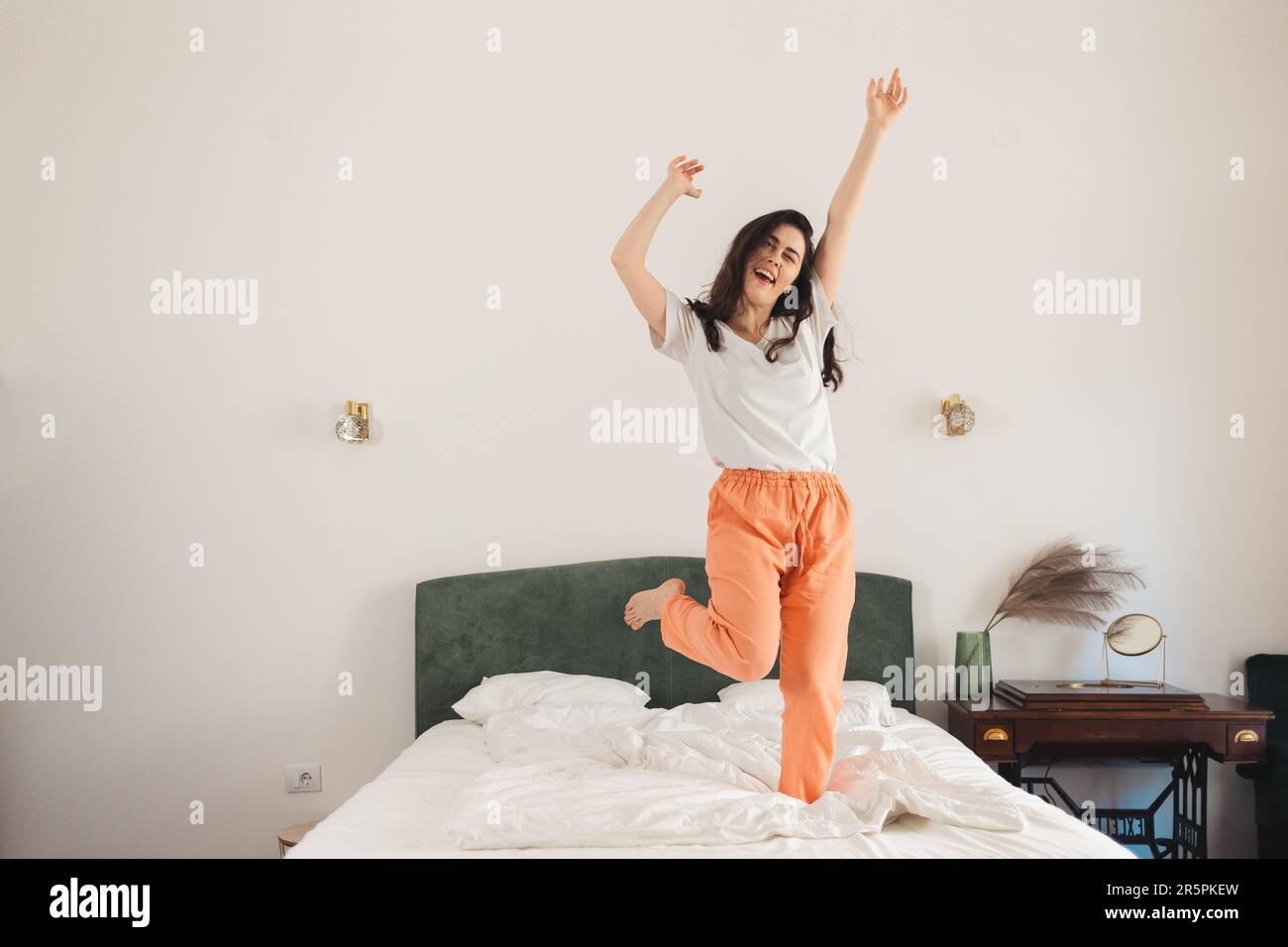 Bonne jeune femme caucasienne danse sur le lit dans la chambre. Liberté et victoire. Banque D'Images