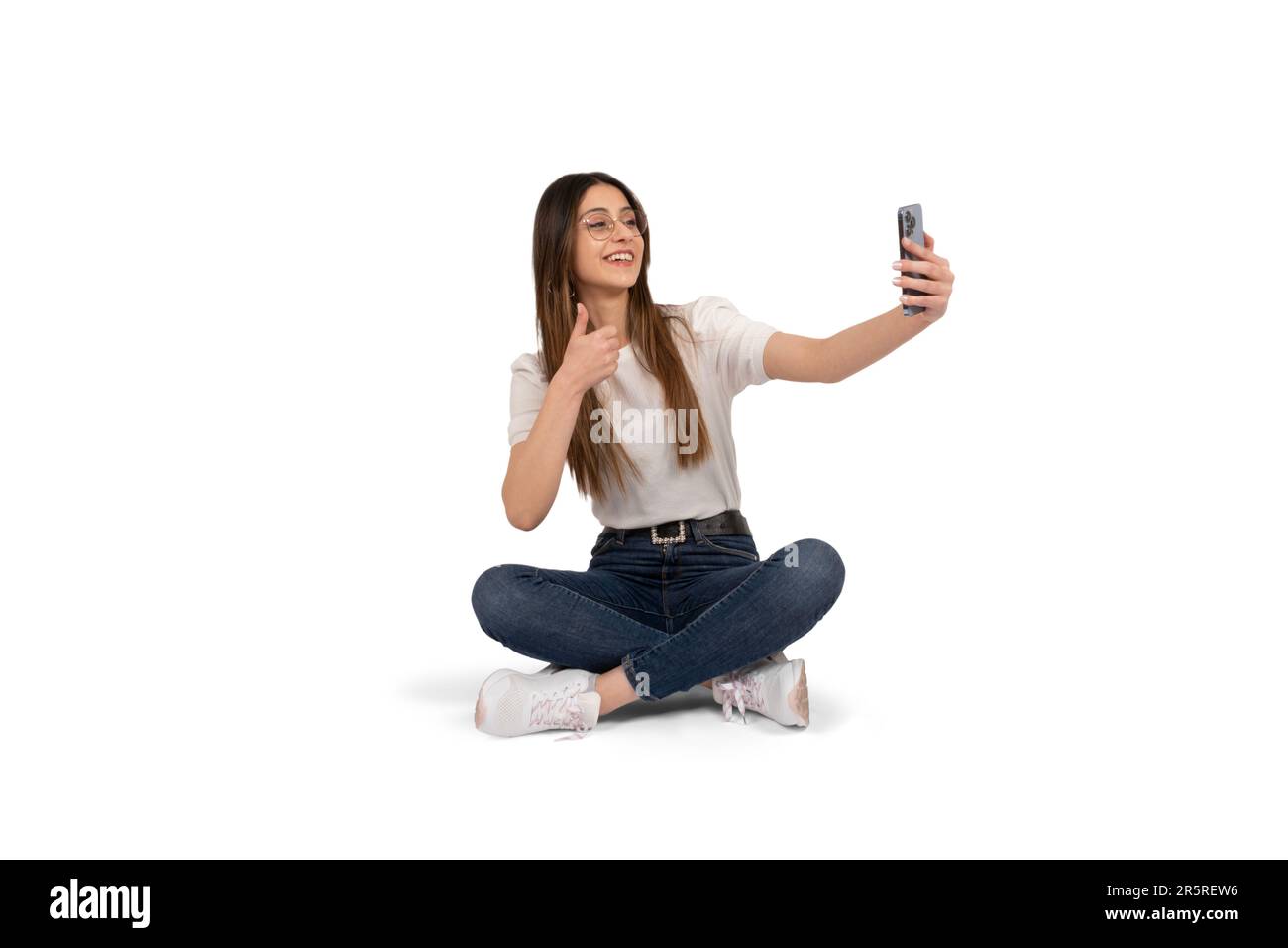 Femme faisant selfie, portrait en pied de jeune femme caucasienne faisant selfie. Assis sur le sol montrant le geste du pouce vers le haut. À l'aide du téléphone portable. Banque D'Images