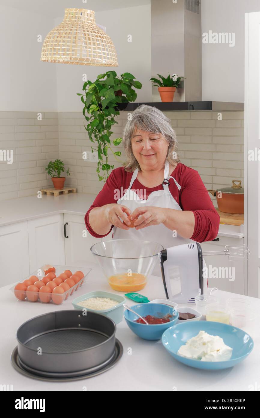 Femme plus âgée à cheveux blancs dans un tablier craquant des oeufs pour faire un gâteau dans sa cuisine Banque D'Images