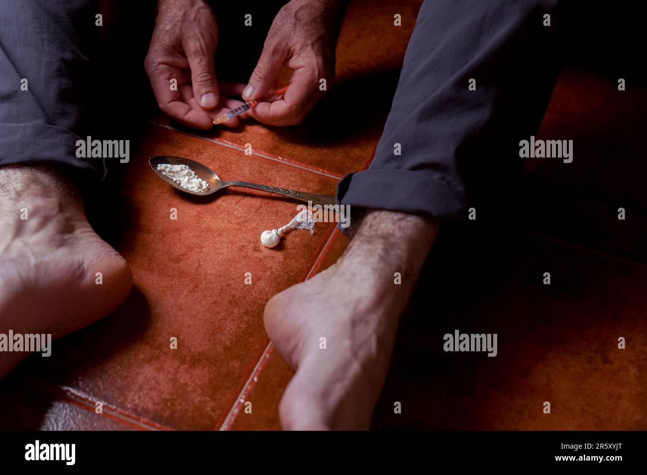 Vue de face d'un homme assis pieds nus sur le sol avec une seringue d'héroïne dans sa main concept de toxicomanie Banque D'Images